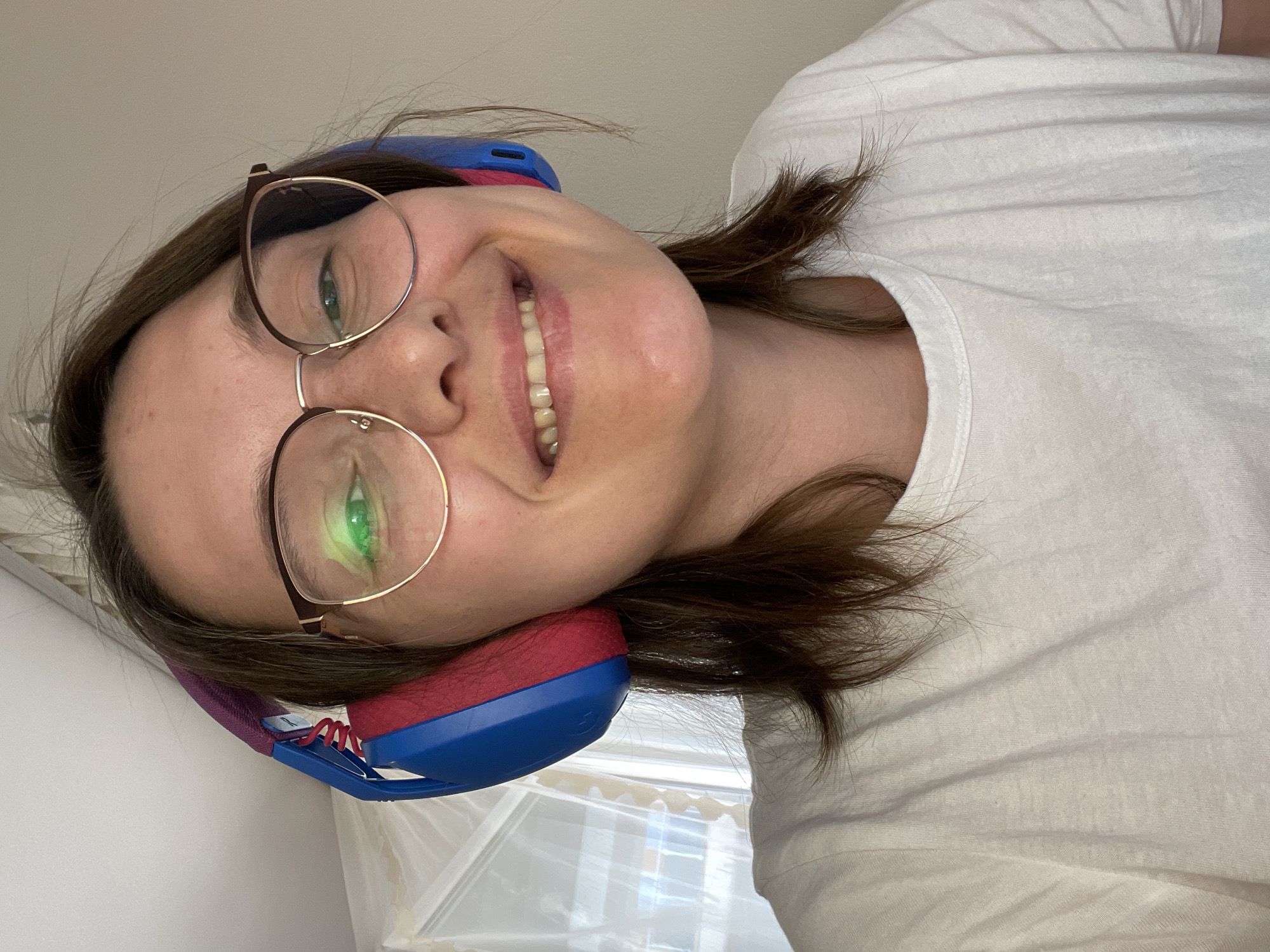 A woman with brown hair and glasses is smiling and wearing blue and red headphones. She is wearing a white t-shirt. The location is not discernible in the image.