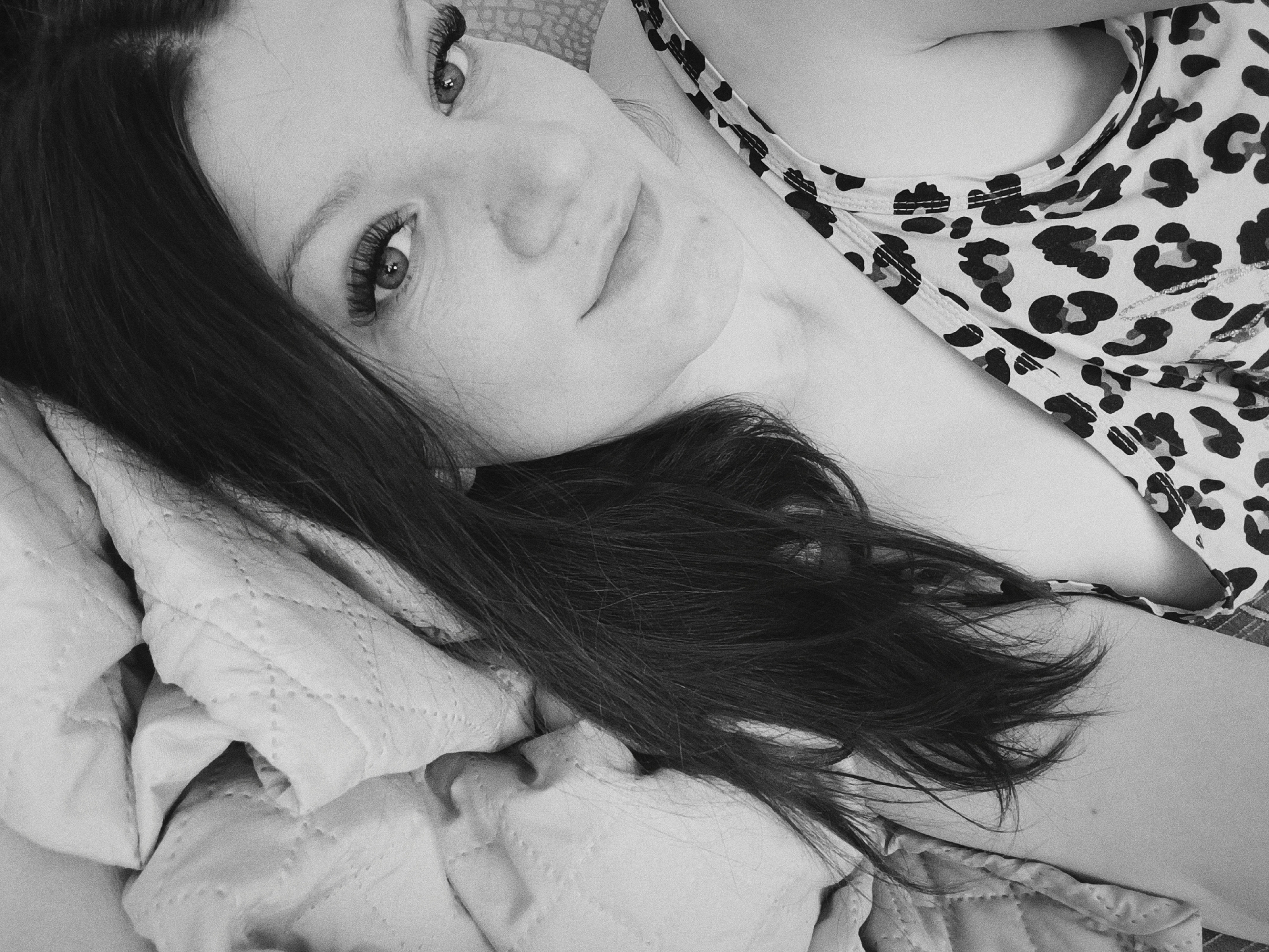 A black and white image depicts a woman with dark hair lying on what appears to be a quilted blanket. She is wearing a top with a leopard print pattern. The woman is looking directly at the camera with a slight smile. The background is indistinct, but the overall setting appears to be indoors. There is no specific information to identify this image as being taken in Tobolsk, Russia.
