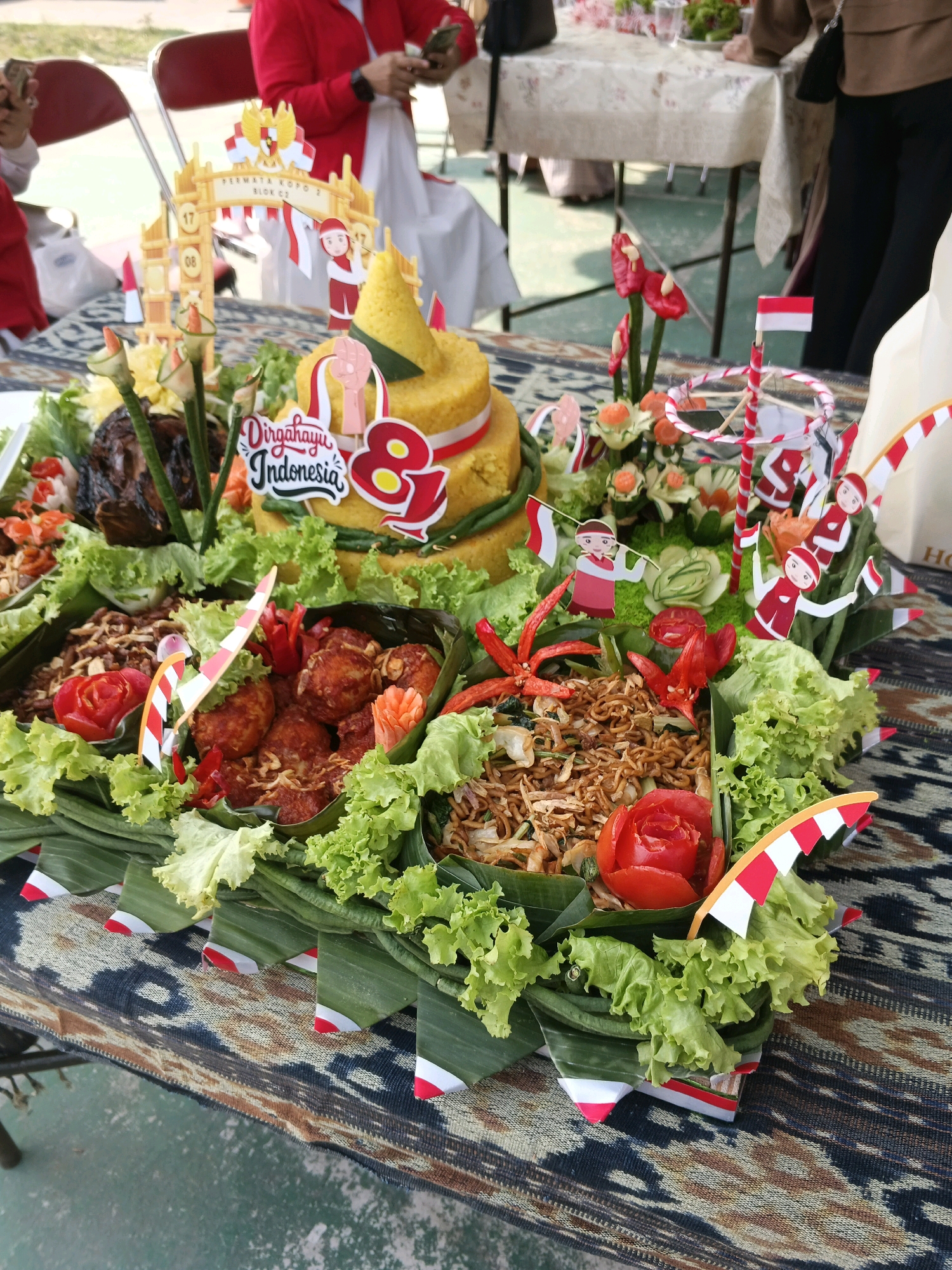 The image shows a close-up view of a festive food display, likely for a celebration in Margahayu, Indonesia. In the foreground, a tiered dish, possibly a Nasi Tumpeng, is decorated with various food items. These include what appears to be fried chicken, noodles, and a large red tomato carved into a rose. The food is arranged on banana leaves and surrounded by lettuce. Several small Indonesian flags and decorative elements featuring cartoon figures holding flags are placed throughout the display.
In the background, a table is visible, laden with more food and drinks. Several people are partially visible, suggesting an event or gathering is taking place. One person, wearing a red jacket, is looking at their phone. Another individual, in the background, is also engaged with a phone. The setting appears to be outdoors, with some sort of pathway or pavement visible on the ground. The overall impression is of a community celebration or event in Indonesia.