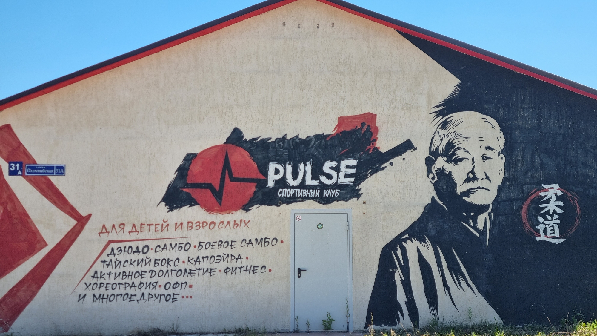 The image shows a mural on the side of a building located at 31A Olimpiyskaya Street in the city of Verzilovo, Russia. The mural features a stylized red heart with a pulse line, the word "PULSE" in large white letters, and "SPORTIVNY KLUB" (sports club) in smaller letters below. To the left of the text, there are red abstract shapes. To the right of the text, there is a black and white portrait of an elderly Asian man in traditional attire, with a circular emblem containing Asian characters next to him. Below the main text, there is a list of activities offered by the sports club, including "DZYUDO," "SAMBO," "BOEVOE SAMBO," "TAISKII BOKS," "KAPOEIRA," "AKTIVNOE DOLGOLETIE," "FITNES," "KHOREOGRAFIYA," "OFP," and "I MNOGOE DRUGOE" (and many more). A white door is visible in the center of the building, behind the mural. The sky is clear and blue.