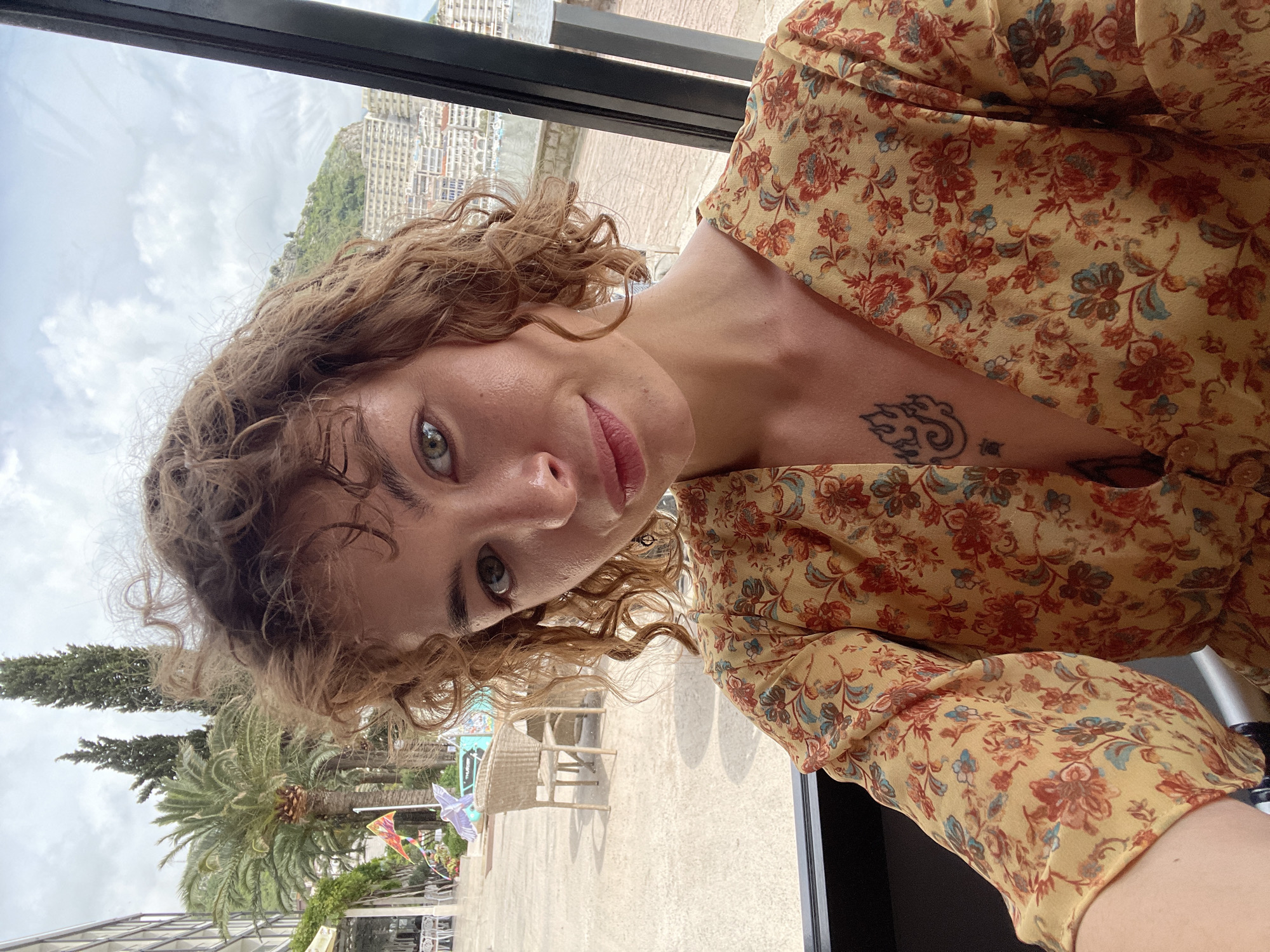 The image is a selfie of a light-skinned woman with curly, light brown hair and green eyes. She's wearing a yellow floral print dress and has a tattoo on her upper chest. The background shows an outdoor scene with palm trees and other greenery, suggesting a tropical or coastal location. Buildings can be seen in the distance, along with a cloudy sky. The location is Rafailovići, Montenegro. The woman appears to be inside, possibly near a window or open doorway, based on the window frame visible in the image. A rattan chair is partially visible outside. The overall impression is a casual, personal snapshot taken in a scenic setting.