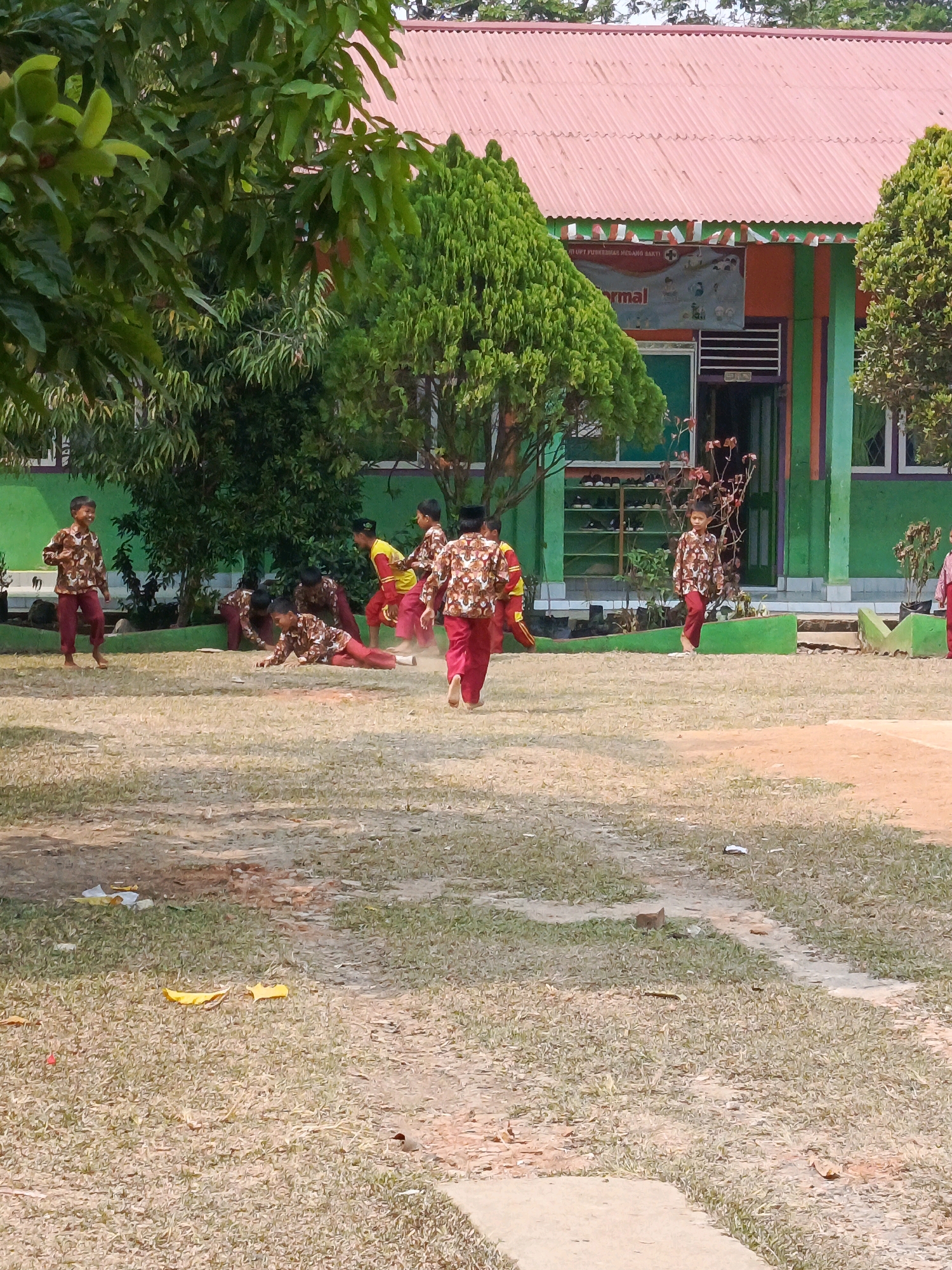 The image depicts an outdoor scene at what appears to be a school or community building in Megang Sakti IV, Indonesia. A group of children, likely students, are engaged in various activities on a grassy field. Several children are wearing matching maroon and white patterned shirts and maroon pants, while one child is in a yellow shirt and red pants. Some children are running, others are interacting, and one appears to be lying on the ground. In the background, there is a green building with a red corrugated metal roof. A banner with text is visible on the side of the building, and trees and foliage surround the area. The ground is covered with dry grass and patches of dirt.