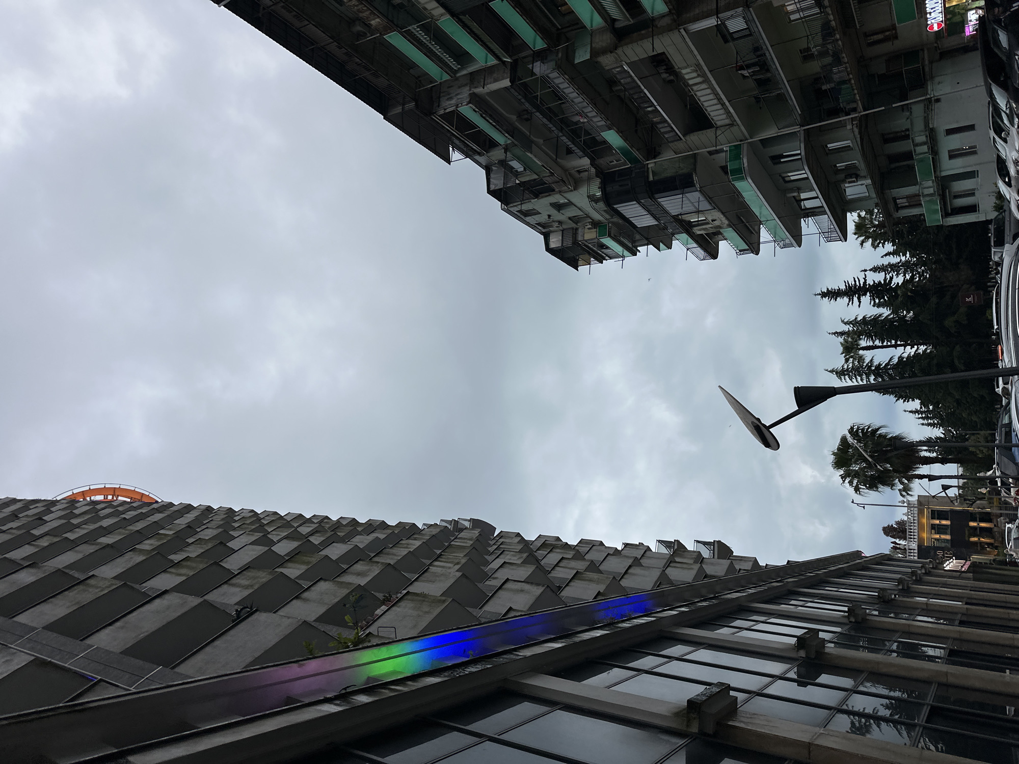 The image is taken from a low angle looking up at buildings and the sky, presumably in an urban area. The bottom building is modern with a series of angled, triangular protrusions along its roofline, as well as large glass windows. A light reflecting colorful light.
The sky is overcast with grey clouds. The top of the image features an older building with a green facade. There are also trees and street lighting visible in the background. Some cars are parked along the street. The building with the triangular roof is likely the BATUMI TOWER.