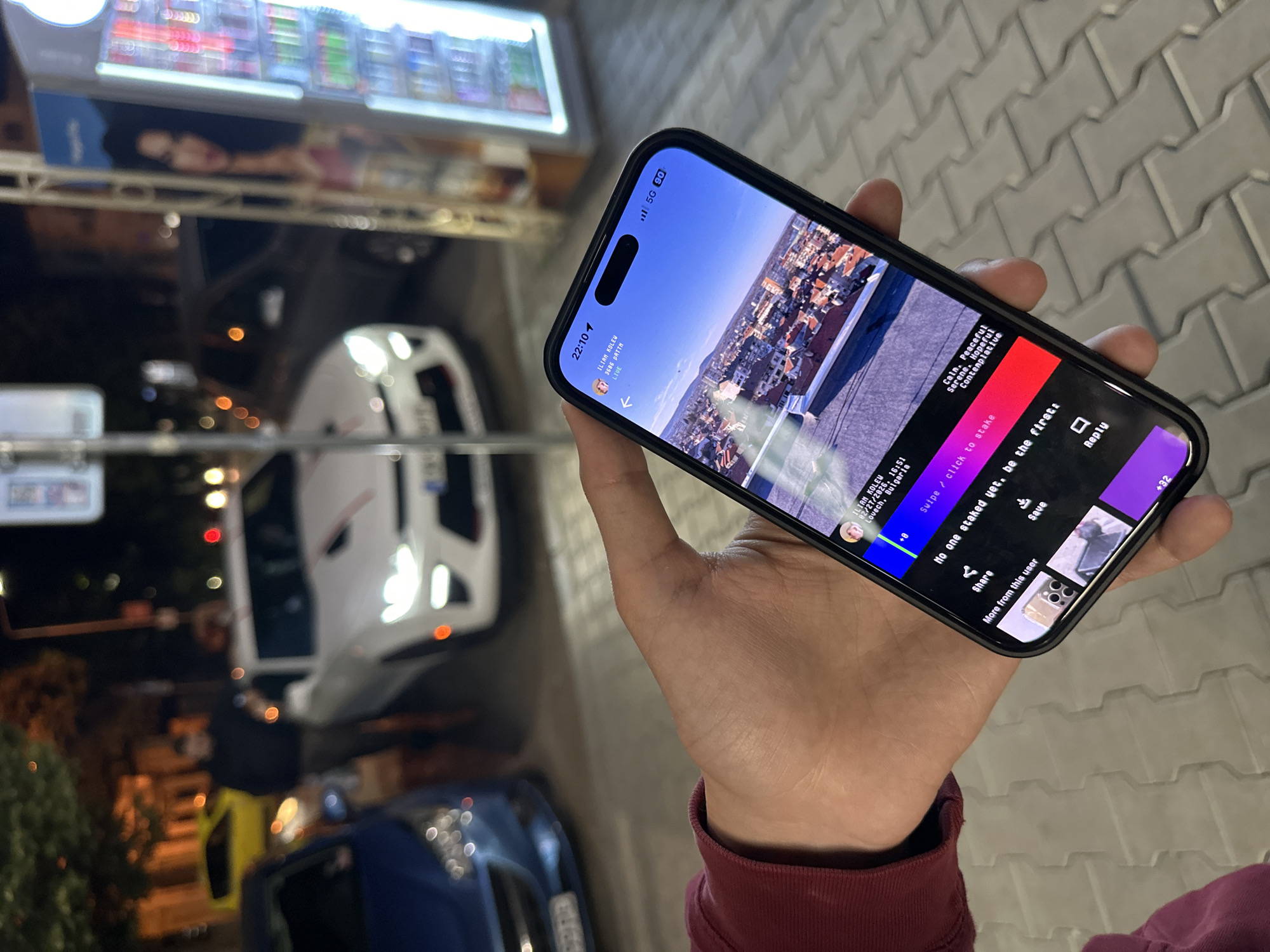 Here is a detailed description of the image:
**Overall Impression:**
The image presents a nighttime scene viewed through the screen of a smartphone held by a person. The phone displays a social media post of a cityscape under a clear sky.
**Smartphone Details:**
The smartphone is black with a notch at the top of the screen. A social media interface is displayed on the screen, which features a live video feed from a user named "ILIAN KOLEW" located in Lovech, Bulgaria. The video features a cityscape, overlaid with text indicating filters and a prompt to stake on the content.
**Foreground Elements:**
A person's hand holds the smartphone. The person is wearing a maroon-colored long-sleeved shirt.
**Background Details:**
The background is blurry and appears to be an urban environment at night. There are multiple cars, including a white van and a blue car. Above the cars, there is a well-lit display of snack food items on a metal rack.
**Location Guess:**
Based on the cityscape displayed on the phone, it could be speculated that the photograph on the phone is showing the city of Veliko Tarnovo. However, the phone claims the user is streaming from Lovech, Bulgaria. Since Lovech is not pictured, we are limited to saying that the phone contains an image of the Veliko Tarnovo landscape.