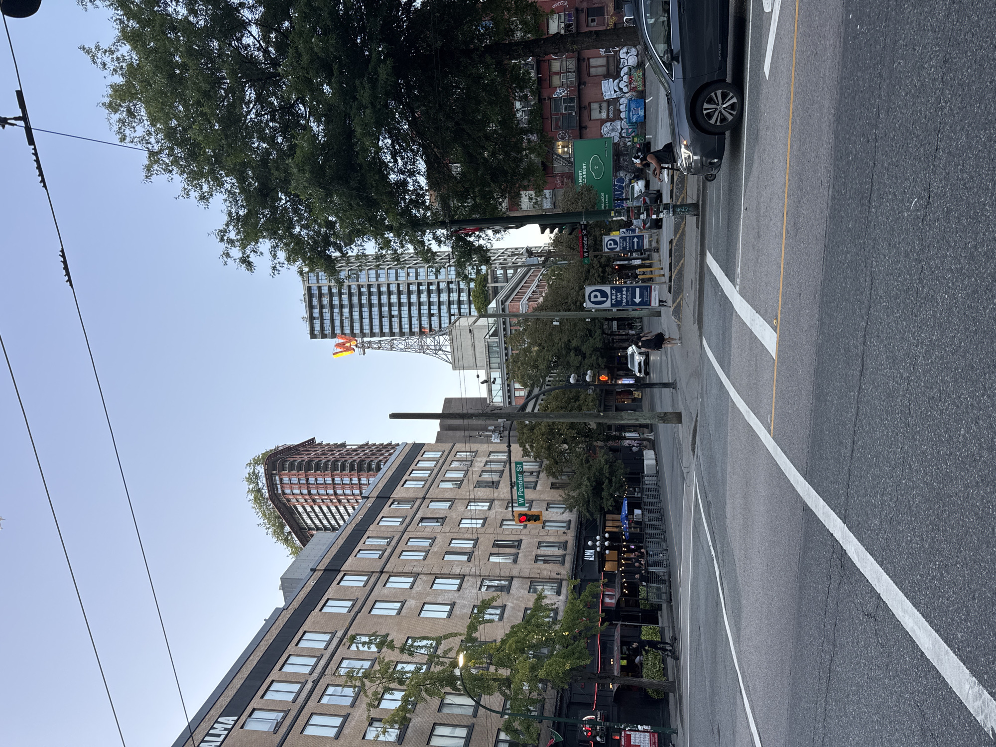 This image captures a street scene in Vancouver, Canada, likely in the Gastown or Chinatown area, identifiable by the distinct architectural styles and the presence of the "W" sign on a tall building. The perspective is from street level, looking across an intersection with visible lane markings and a traffic signal.
On the left side of the street, a multi-story brick building with numerous windows is prominent, with signage indicating "ALMA" on the ground floor. Trees line the sidewalk in front of the building, providing some greenery. Further down the street, other buildings with various architectural features are visible, including a tall, modern skyscraper topped with a large, illuminated "W" sign.
On the right side of the intersection, a dark-colored SUV is the most prominent vehicle, positioned in the foreground. Other vehicles are present further down the street. Two parking signs are visible, indicating "PUBLIC PAY PARKING." Pedestrians are also present, including a person on a scooter near the left sidewalk and a few individuals walking on the right side of the street. The sky is clear and blue, suggesting it's daytime. The overall atmosphere appears to be that of a busy urban environment with a mix of historical and modern architecture.