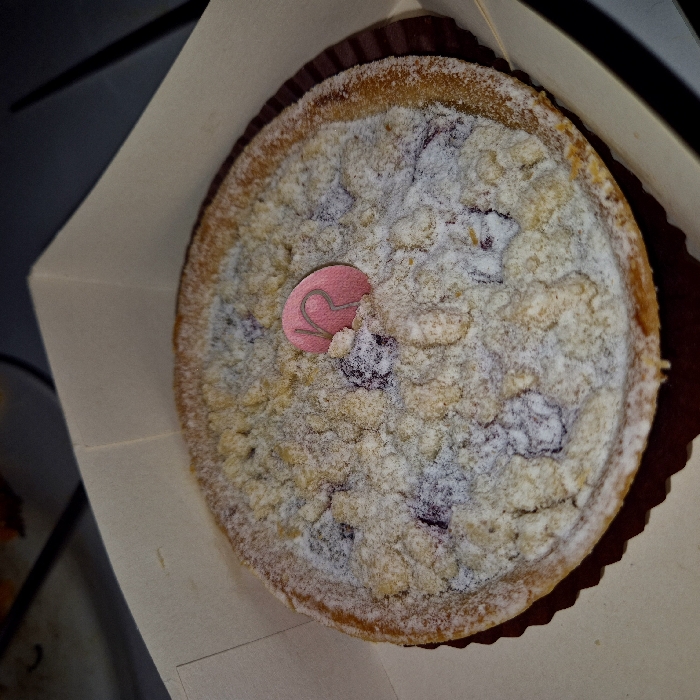 A single, circular crumble tart is depicted within an open, light beige corrugated cardboard takeaway box. The tart rests in a dark brown, fluted paper liner. Its surface is covered with a coarse, light-colored crumble topping, heavily dusted with white powdered sugar. Dark berries, consistent with blackberries or blueberries, are partially visible beneath the crumble. A small, oval-shaped pink tag, bearing a stylized capital "R," is inserted vertically into the crumble topping slightly off-center to the right. The visible edge of the tart's crust is golden-brown. The interior walls of the takeaway box frame the pastry. The background beyond the box is dark and out of focus, providing no additional environmental details or visual evidence of people, actions, or interactions. Based on the distinctive "R" branding, the pastry originates from Pâtisserie Régis, a known establishment in Strasbourg, France.
