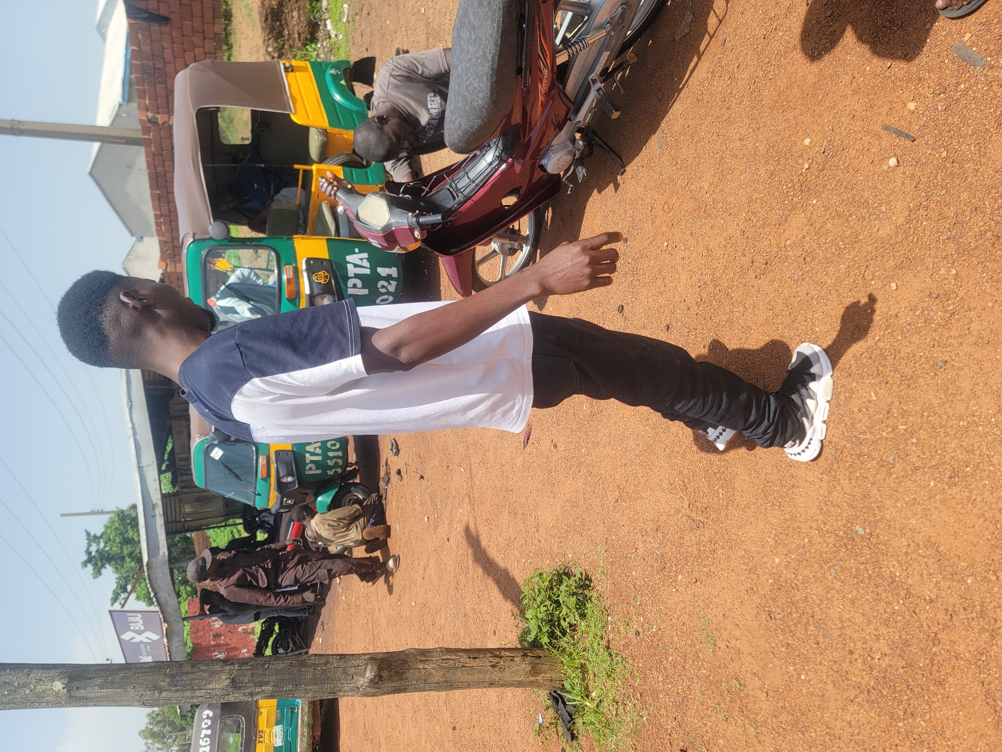 In Barakin Lerawi, Nigeria, an outdoor scene unfolds with a group of individuals interacting around vehicles. The ground is a reddish-brown dirt surface, scattered with small stones and debris. In the background, several colorful auto-rickshaws, also known as "keke napep," are parked. One of these rickshaws, predominantly green and yellow, displays "PTA - 0510" on its side. Another auto-rickshaw, partially visible on the left, is yellow with green accents and has the number "6799" on it.
A maroon motorcycle is a central object, with a man in a grey t-shirt leaning over it, seemingly working on the engine. He appears focused on his task, with his arms beneath the motorcycle.
A man in a dark brown outfit, including a cap and trousers, stands near the auto-rickshaws, observing the scene. His hands are not clearly visible.
In the foreground, a person, seen from the waist down, is walking away from the camera. They are wearing dark pants and white and black athletic shoes with a distinctive white sole pattern. Their shadow stretches out on the ground.
Another individual, whose upper body is visible from the back, stands to the right of the frame, also looking towards the motorcycle and the people around it. This person is wearing a white t-shirt with dark blue trim around the collar and shoulders.
The environment suggests a somewhat dusty and unpaved area, possibly a roadside stop or a local transport hub, with some vegetation growing near a wooden post on the left. The sky is bright and clear, indicating a sunny day. The overall impression is one of everyday activity related to transportation and mechanics in a Nigerian setting.