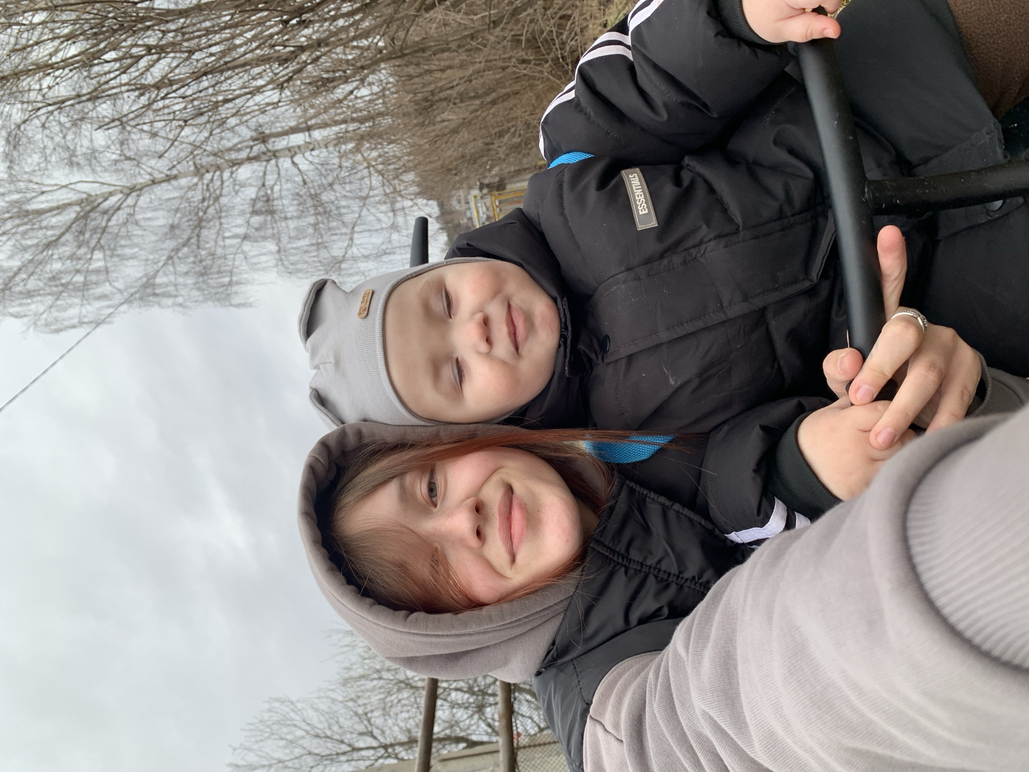 A mother and child are captured in a selfie-style photograph taken in an outdoor environment, likely in the city of Фоки, Russia. The child, wearing a gray knit hat and a black puffy jacket with the word "Essentials" on the left breast area, is snuggled close to the mother. The mother is wearing a gray hooded sweatshirt and a black puffy vest. They are holding onto a black metal bar, possibly part of a swing or playground equipment. The background features bare trees against an overcast sky.