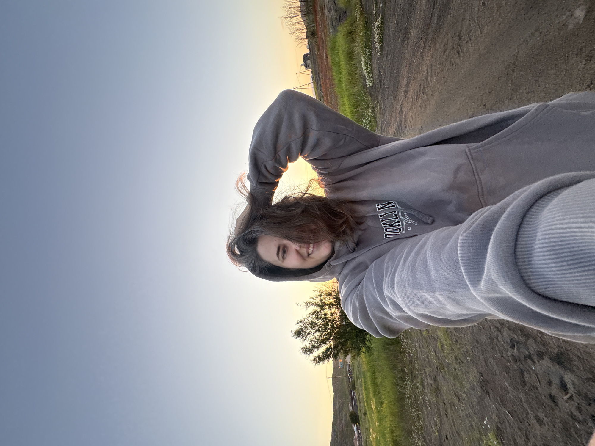 A selfie taken from a low angle shows a young woman with dark hair, wearing a grey hooded sweatshirt. She is holding her hair up with her right hand, which is near her head, and her left arm is extended towards the camera. Her face is visible, and she is smiling. The background depicts a rural or undeveloped area with sparse vegetation, a dirt road, and a few distant buildings. The sky is a clear gradient of pale blue to yellow, suggesting it is either sunrise or sunset. The overall lighting is soft and warm, characteristic of a golden hour. The environment appears to be the outskirts of Teriberka, Russia, based on the landscape.