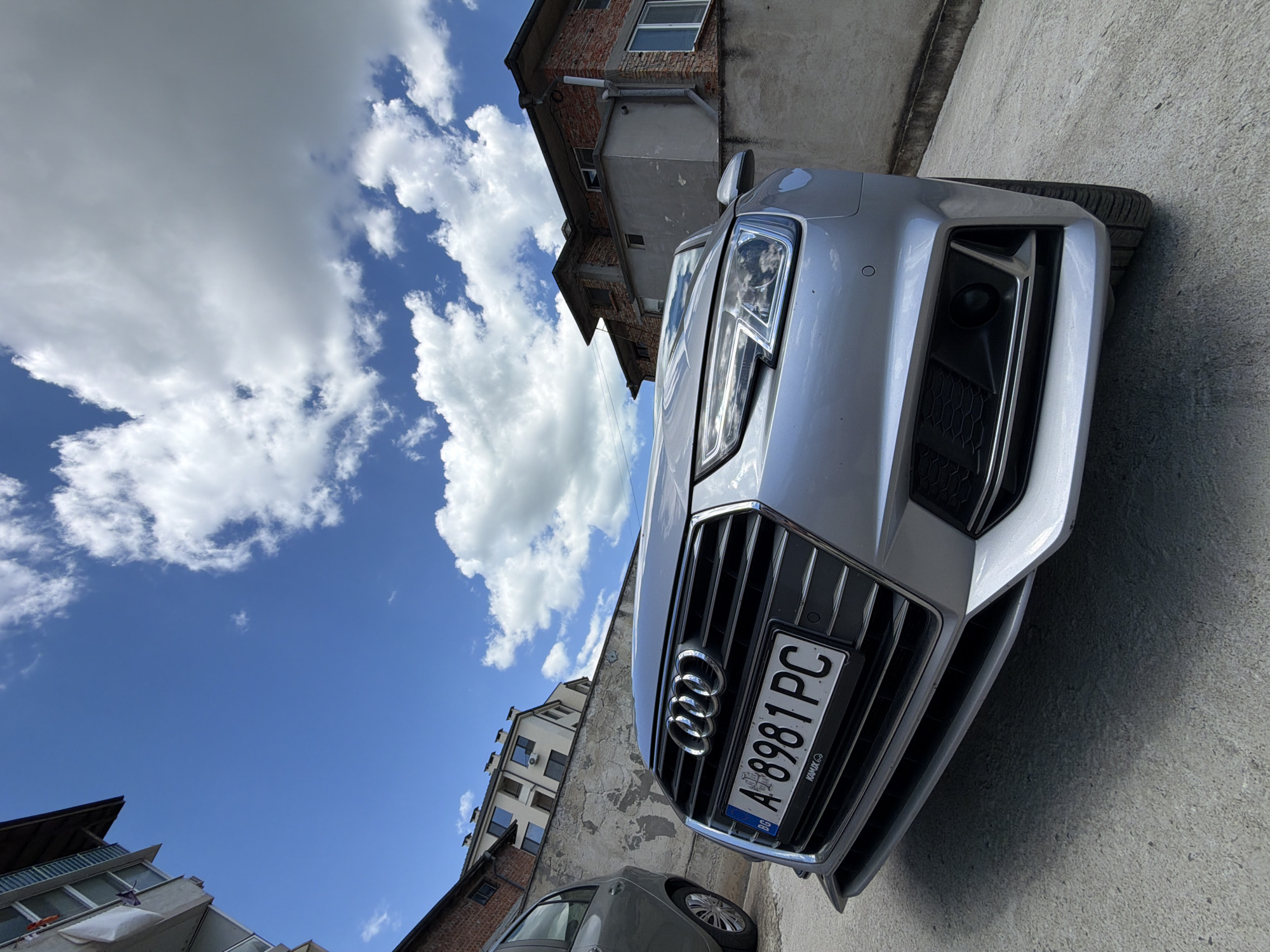 An Audi A4, license plate A 8981 PC, is parked on a concrete surface outdoors. The car is silver and is angled towards the viewer. The foreground shows the front bumper, grille, and headlights of the car, with the license plate clearly visible. Behind the car, a textured concrete wall runs horizontally across the frame. A portion of another car, grey in color, is visible to the left of the Audi. In the background, there are multi-story buildings constructed with brick and concrete, under a blue sky with scattered white clouds. The scene appears to be in a residential or urban area.
