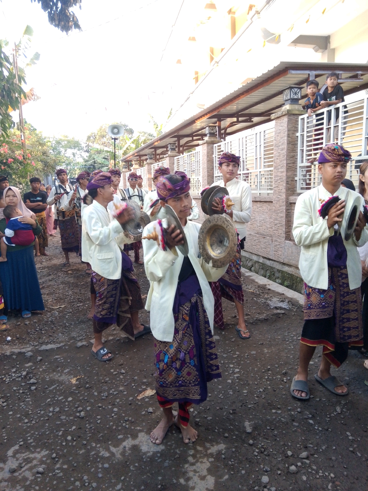 The image depicts a street scene in Kuripan Utara, Indonesia, during what appears to be a cultural procession. Several young men, dressed in traditional Balinese attire featuring sarongs and headbands, are walking in a line. They are holding and playing musical instruments that resemble cymbals or gongs. The environment is an unpaved street with gravel and some wet patches. In the background, there is a building with a corrugated metal roof and decorative railings. A few spectators, including a woman holding a baby and some children watching from a balcony, are also visible. The overall atmosphere suggests a community event or celebration.