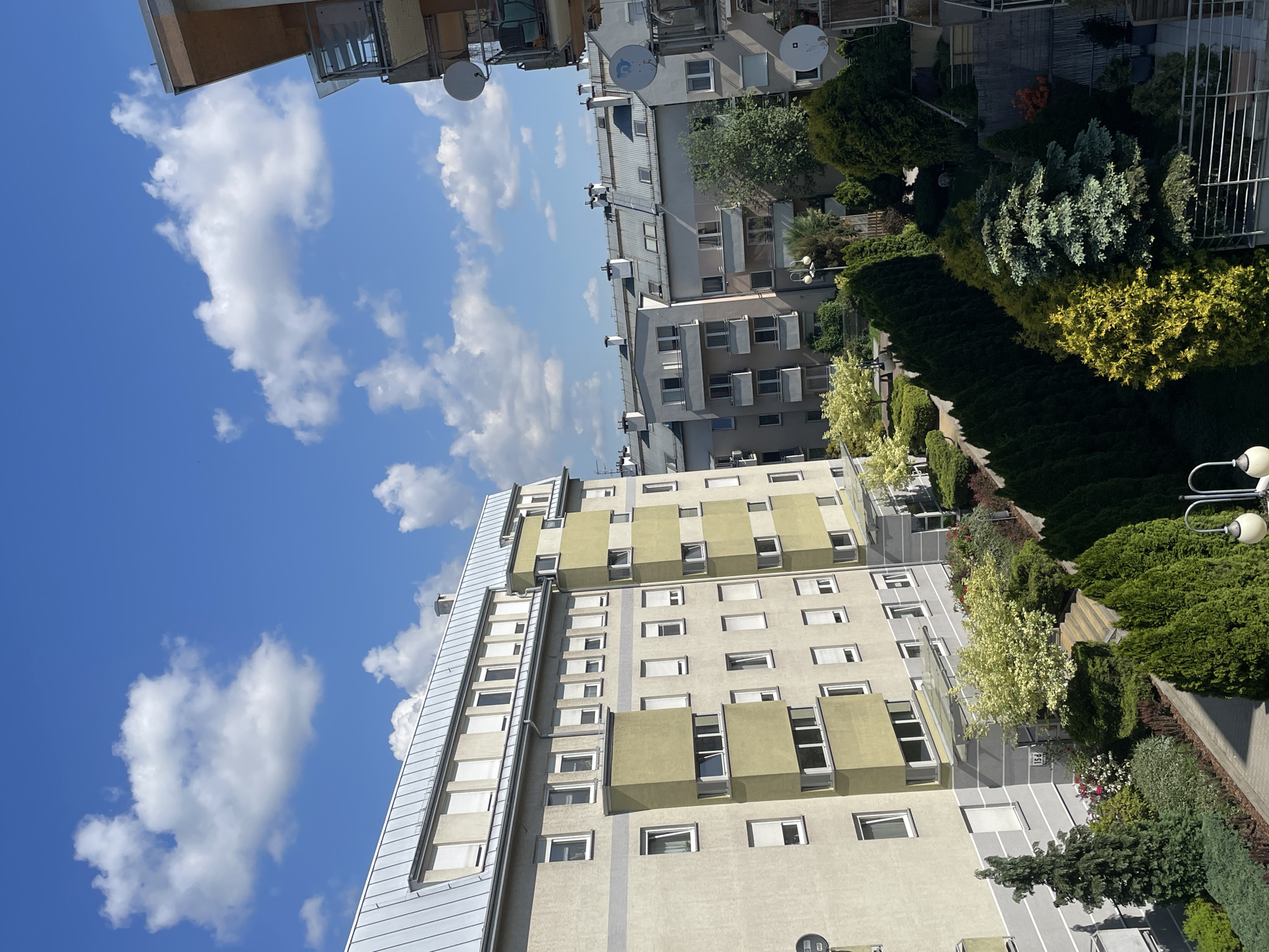 The image shows an aerial view of buildings and green spaces in an urban environment, specifically in Unknown, Unknown. A predominantly white and light gray building with green accents and many windows is visible on the lower half of the image. A light gray building with a metal roof is attached to it. Another building, gray in color, can be seen further away. The sky is blue with scattered white clouds. Dense greenery, including trees and bushes, is present around the buildings. The overall setting suggests a densely populated urban area with a mix of residential or commercial buildings and green landscaping. The sky is visible and it appears to be daytime.