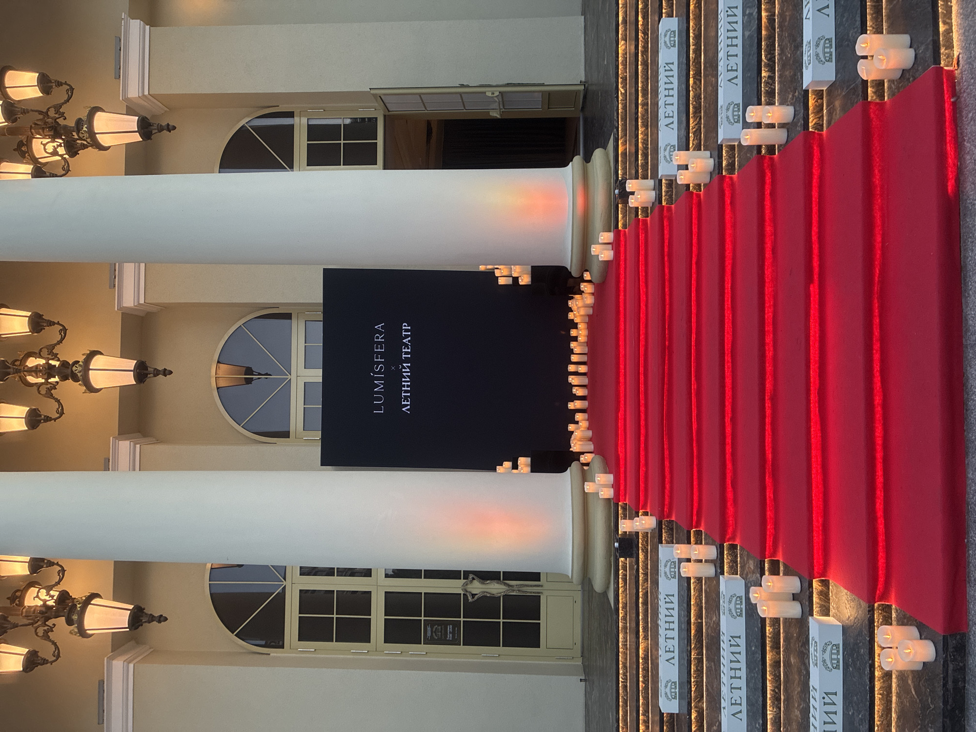 The image depicts the entrance to the "LUMISFERA ЛЕТНИЙ ТЕАТР" (LUMISFERA SUMMER THEATER) in Sochi, Russia. A wide, vibrant red carpeted staircase leads up to a black backdrop displaying the event's name. The staircase is illuminated from below with red lighting, and numerous white pillar candles are strategically placed along its edges and on the steps.
Flanking the carpeted entrance are two large, white classical columns. Above these columns, ornate, vintage-style chandeliers with multiple light fixtures hang, casting a warm glow. Behind the columns and to the sides of the backdrop are large, arched windows with segmented panes. Open doorways can be seen on either side of the building, suggesting an interior space.
The steps leading up to the main entrance are also decorated with white boxes featuring the text "ЛЕТНИЙ" (SUMMER) and an image of a crown or ornate crest. More pillar candles are placed on these steps. The overall scene is formal and festive, suggesting an event or premiere. The ground in front of the staircase appears to be paved.