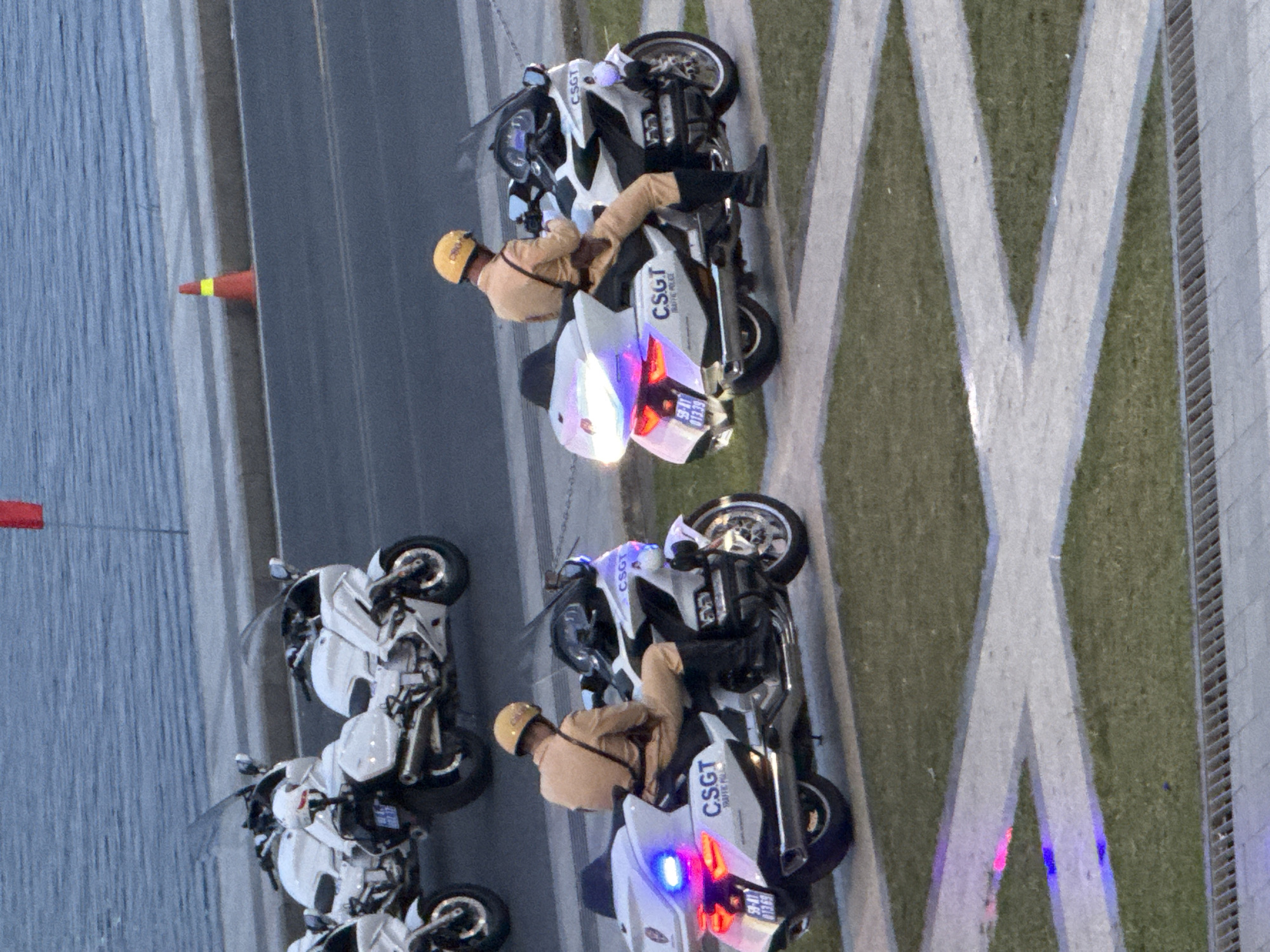 Two police officers in tan uniforms and yellow helmets are seated on white motorcycles with "CSGT TRAFFIC POLICE" markings. The motorcycle on the right has flashing blue and red lights on its rear. The officers appear to be on duty, possibly in a city setting. In the background, a body of water and a traffic cone are visible. This scene is likely in Da Lat, Vietnam, given the presence of the CSGT (Cảnh sát Giao thông - Traffic Police), a Vietnamese law enforcement agency.