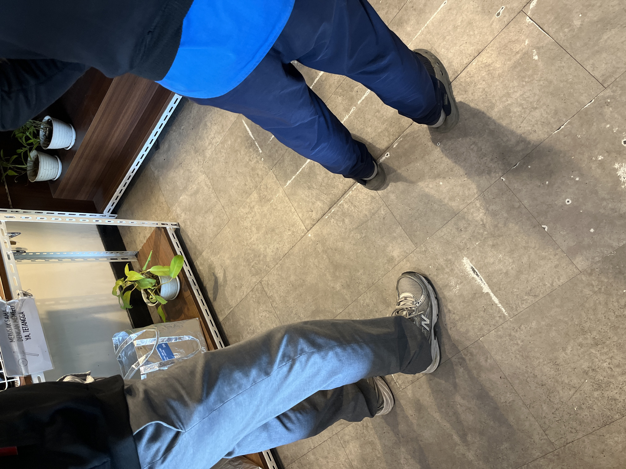 Two individuals, identified only by their lower bodies and legs, are standing in a well-lit indoor space. The floor is tiled in a neutral gray color with visible white scuff marks and some debris. To the left, one person wears gray trousers and silver athletic shoes with a prominent "N" logo. To the right, the other person wears dark blue trousers and dark gray athletic shoes.
In the background, a white metal shelving unit stands against a light-colored wall. On one of the shelves, a small potted plant with green leaves sits next to a translucent, silvery shopping bag. Above the plant and bag, a sign is attached to the shelf, featuring Indonesian text that appears to be a warning or instruction. Further back on another shelf of the same unit, two small potted plants are visible.
The overall impression is that of a retail or storage area, possibly a shop or a stockroom. The presence of shelving, plants, and a sign suggests a functional commercial environment. The image captures a moment in time, with no overt actions or interactions taking place between the individuals, who are positioned some distance apart. The scene appears to be clean and organized, apart from the floor markings.