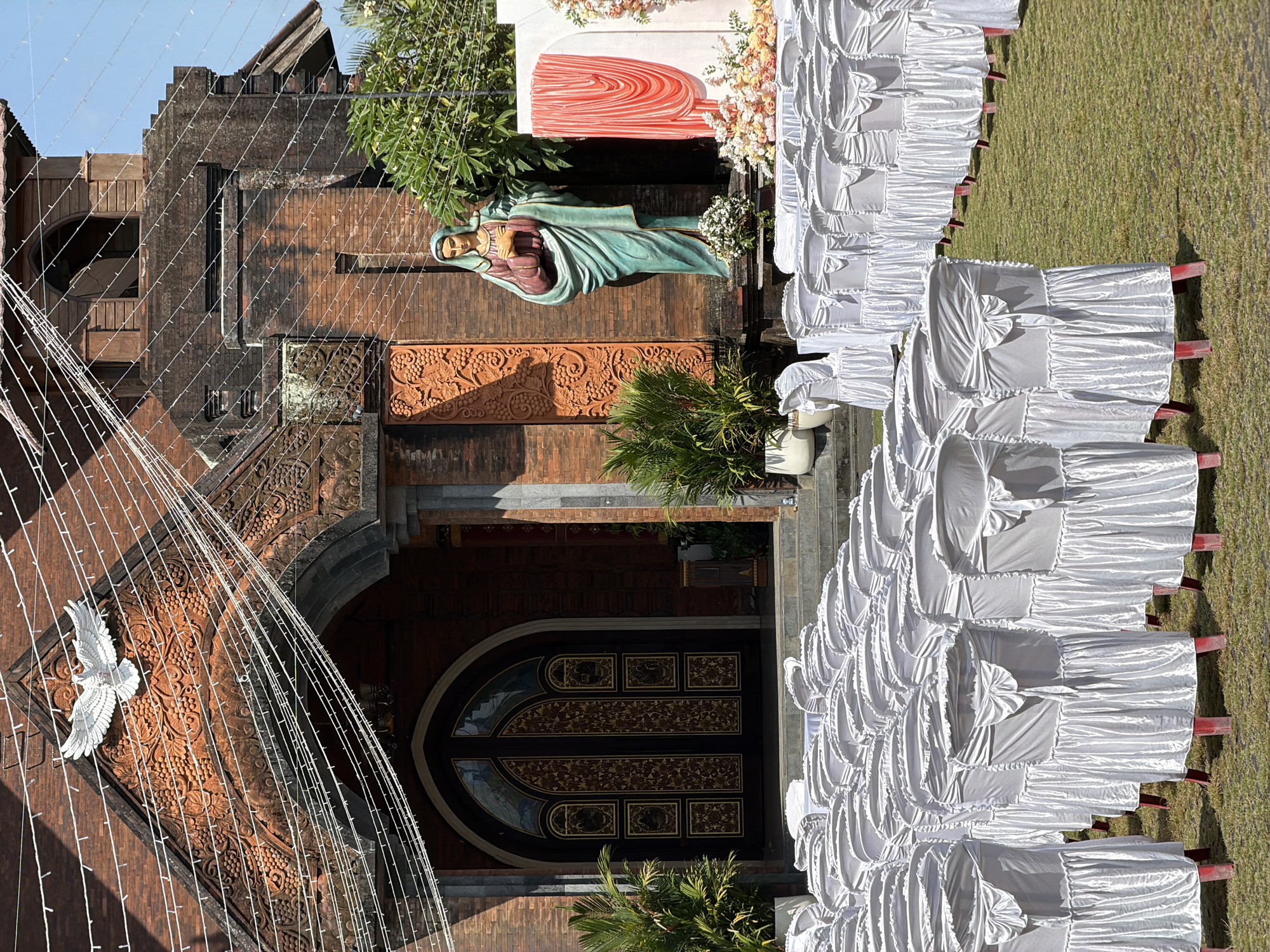 A wide shot captures an outdoor wedding ceremony setup in front of a brick church in North Kuta, Indonesia. The church features ornate carvings, a large arched doorway with decorative panels, and a white dove sculpture above the entrance, all adorned with strings of white fairy lights. To the right of the church, a statue of a robed figure stands near a backdrop of coral-colored drapes and floral arrangements. The foreground is dominated by rows of white-covered chairs, neatly arranged on a grassy lawn, awaiting guests. The sky is clear and blue.