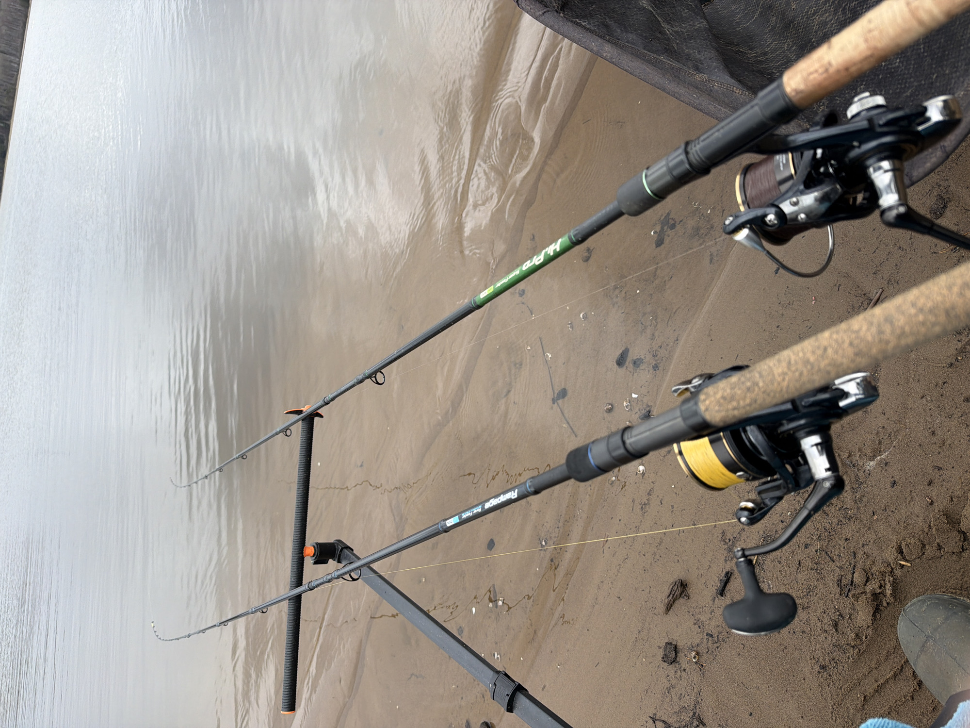 Here's a description of the image:
Two fishing rods are set up on a sandbank, facing a body of water. The rods are supported by a rod stand. The rod closer to the viewer is a "Rampage" rod. A black and silver fishing reel with yellow line is attached to it. The second rod, further away, is a "H-Pro" rod. Its fishing reel is black and silver with a brown line. The water is calm and reflects the sky. A portion of a dark net is visible in the upper right corner. A boot is visible in the lower right corner. The sand is damp and has some debris on it. The location is likely Chaikovsky, Russia, based on the prompt.