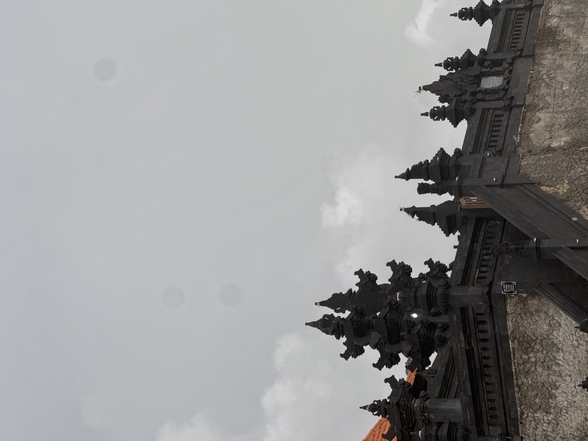An ornate, dark stone temple gate, characteristic of Balinese architecture, dominates the right side of the frame, set against a vast, light grey, overcast sky. The structure is intricately carved with multiple pointed spires, tiered roofs, and detailed relief work, appearing to be crafted from dark volcanic stone.
Small, lighter-colored objects, possibly offerings or decorations, are perched on several spires. A small rectangular sign is affixed to the dark stone, bearing the English text "ONLY ENTRY" and the name "PURA KEHEN." Below "ONLY ENTRY," some Indonesian words, likely "PINTU MASUK" (Entrance), are also visible. An adjacent wall to the right of the gate is constructed from a lighter, textured stone, and a glimpse of an orange-tiled roof is visible at the bottom edge.
The sky is dull and uniform, with faint white clouds, suggesting an overcast or cloudy daytime. Several soft, circular grey smudges are visible in the sky, indicative of lens flares or sensor dust. The scene captures the grandeur of the temple entrance in what appears to be North Kuta, Indonesia, under a muted sky.
