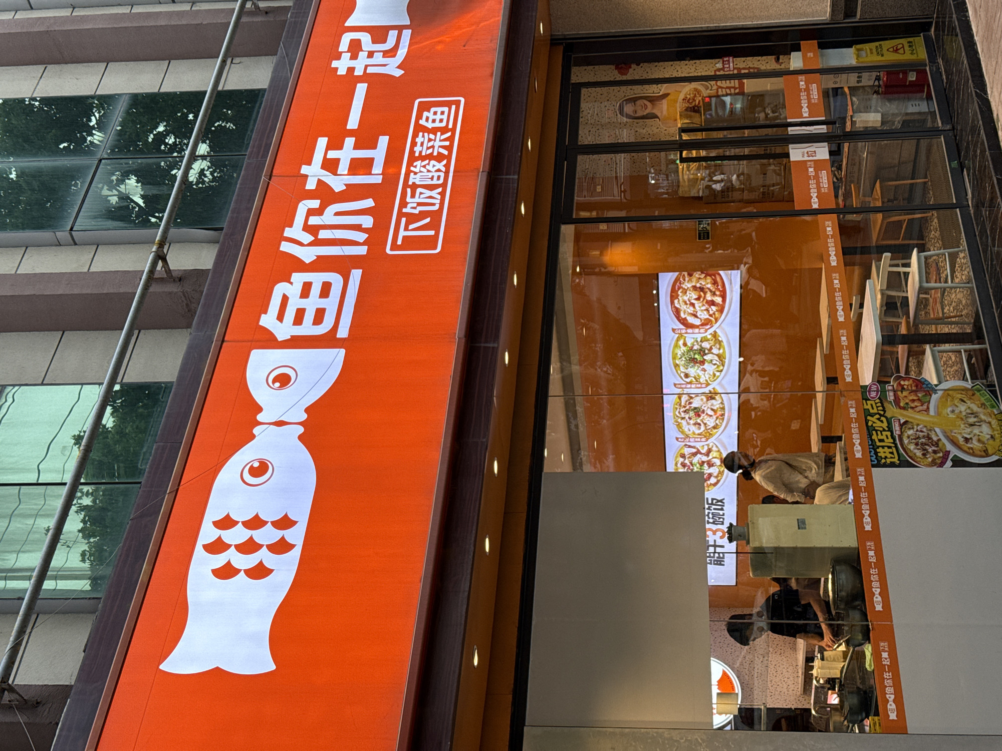 The image shows the exterior of a restaurant called "Fish You're Together" (鱼你在一起) in Lintong District, China. The orange sign features a white fish silhouette and Chinese characters. The restaurant's interior is visible through the glass windows, revealing customers dining, hanging food posters, chairs and tables. Reflections can be seen on the glass, including images of pedestrians and traffic. Additional signage is along the edges of the windows.