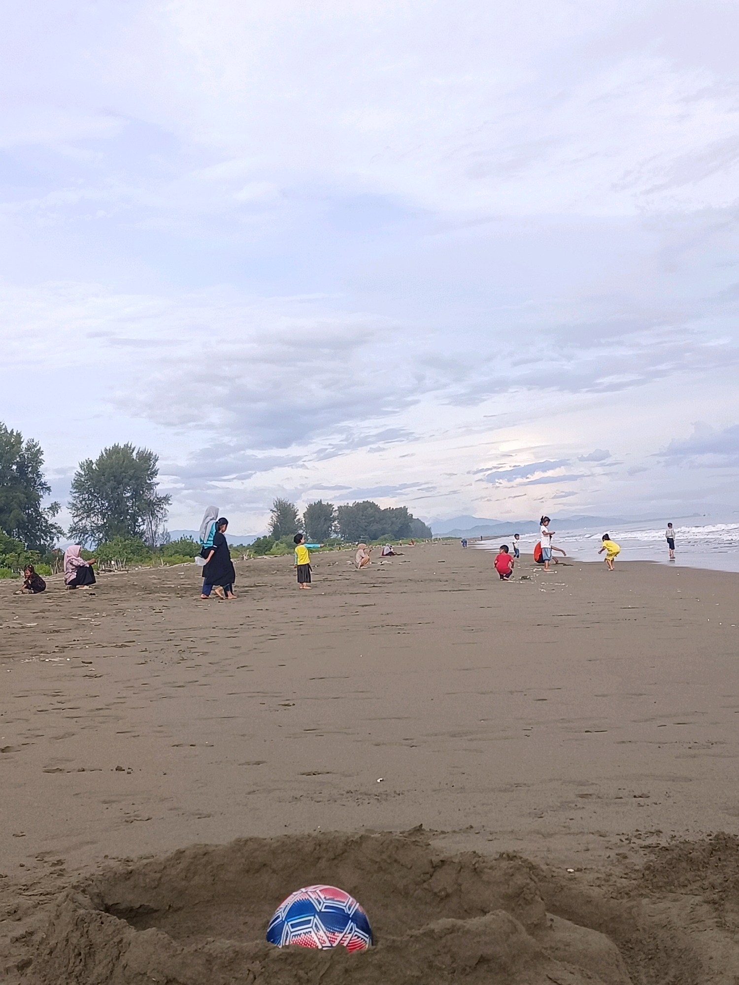 The image depicts a cloudy day at a sandy beach in Reudeuep, Indonesia. In the foreground, a partially buried soccer ball with red, white, and blue geometric patterns rests in a sand pit. The beach is mostly empty except for a few scattered individuals and small groups.
To the left, closer to the trees, several people are sitting or squatting on the sand. Two women, one wearing a blue headscarf and the other a black hijab and dress, are walking together. Further to the right, a young boy in a yellow shirt and dark shorts stands on the sand.
In the mid-ground, a larger group of people, mostly children, are scattered near the water's edge. A child in a red shirt is crouching, and another in a yellow shirt is mid-jump. Further down the beach, a few more individuals are visible, some standing and others appearing to be sitting or playing. The ocean waves are visible in the background, and the shoreline stretches towards the right. Trees and vegetation line the edge of the beach, with a range of mountains visible in the distance under the overcast sky. The sand appears dark and wet in some areas closer to the water.