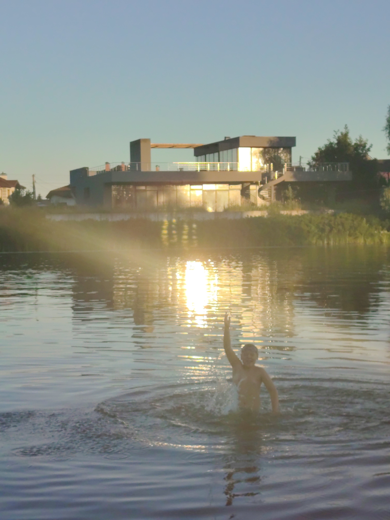 In an outdoor setting, a young boy is shown emerging from the water of a lake or river. He is partially submerged, with his torso and head visible. One arm is raised above his head, and water is splashing around him, indicating he is moving or has just made a splash. The sun is low in the sky, casting a bright reflection on the water and creating lens flares across the scene. In the background, a modern, multi-story building with large windows and a flat roof is visible, situated on the shore. Trees and other vegetation can be seen behind the building, and a clear, light blue sky fills the upper portion of the frame. The overall impression is a sunny, relaxed outdoor scene, likely during sunset.