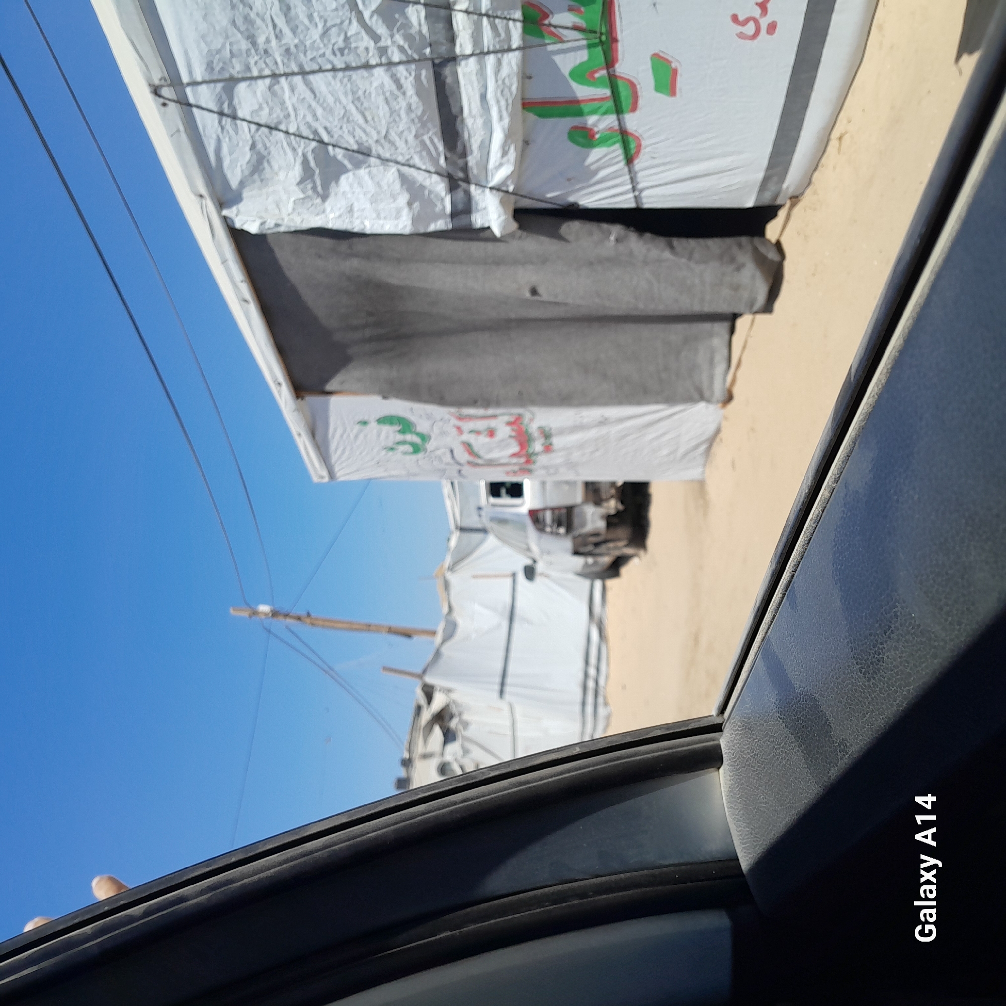 The image, taken from inside a vehicle, shows a view of a camp-like setting in Deir el-Balah, Palestinian Territories. In the foreground, the black interior of the car door and window frame are visible, with the text "Galaxy A14" in the lower left corner. Beyond the car, a row of white tents or temporary structures stretches across the frame. A light-colored, older model car with its rear bumper removed is parked amongst these structures. One of the tents has Arabic writing in green and red. Power lines are visible against a clear blue sky. The ground is sandy.