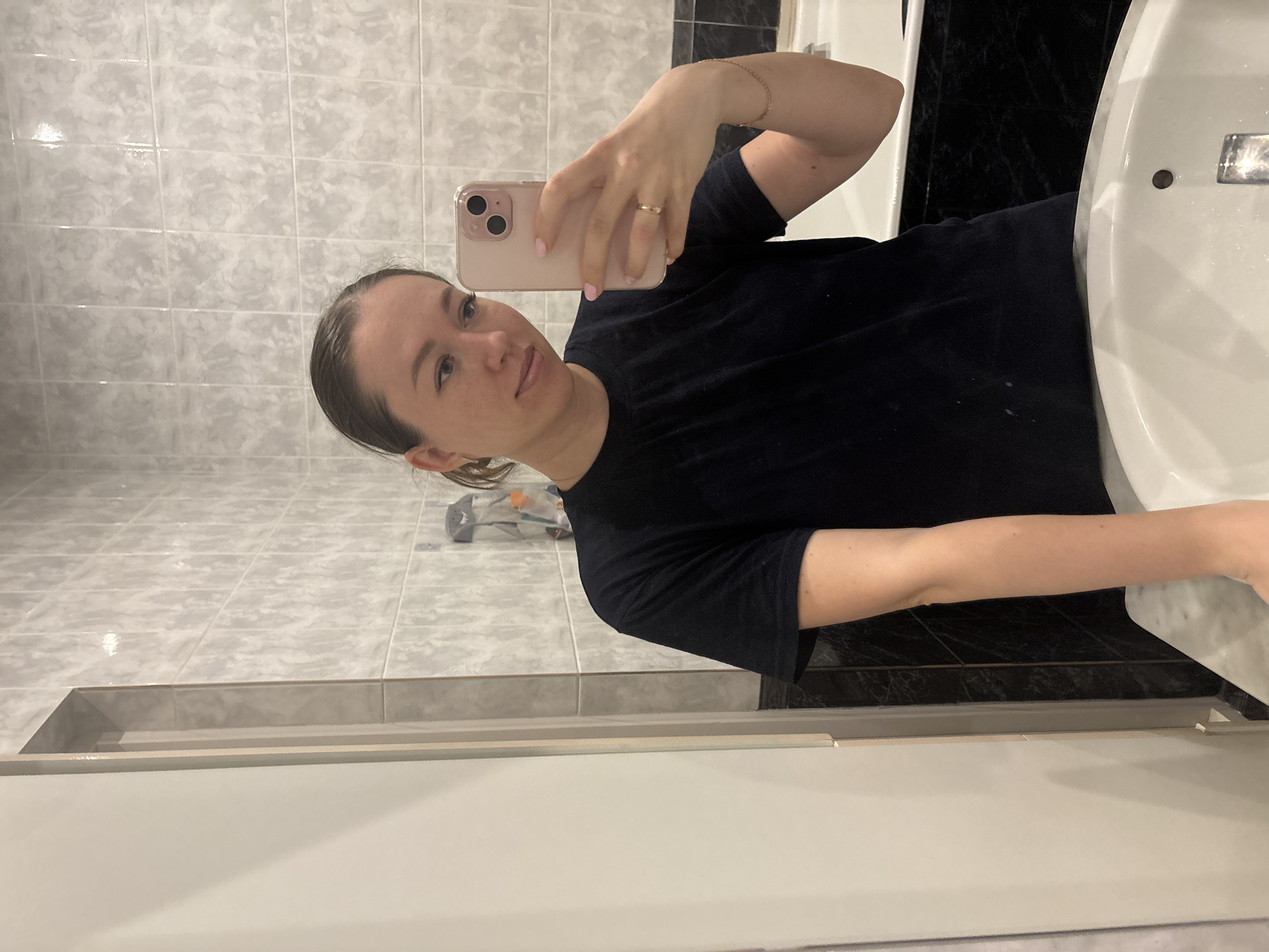 A woman is taking a selfie in a bathroom. She's holding a light pink phone with two rear-facing cameras. She is wearing a black t-shirt and a gold bracelet. Her hair is pulled back, and she has a gold ring on her finger. The bathroom features light gray tiled walls, a white sink, and black marble-like tile behind the sink. A toothbrush holder is visible near the sink. The location is identified as Ферма, Russia.
