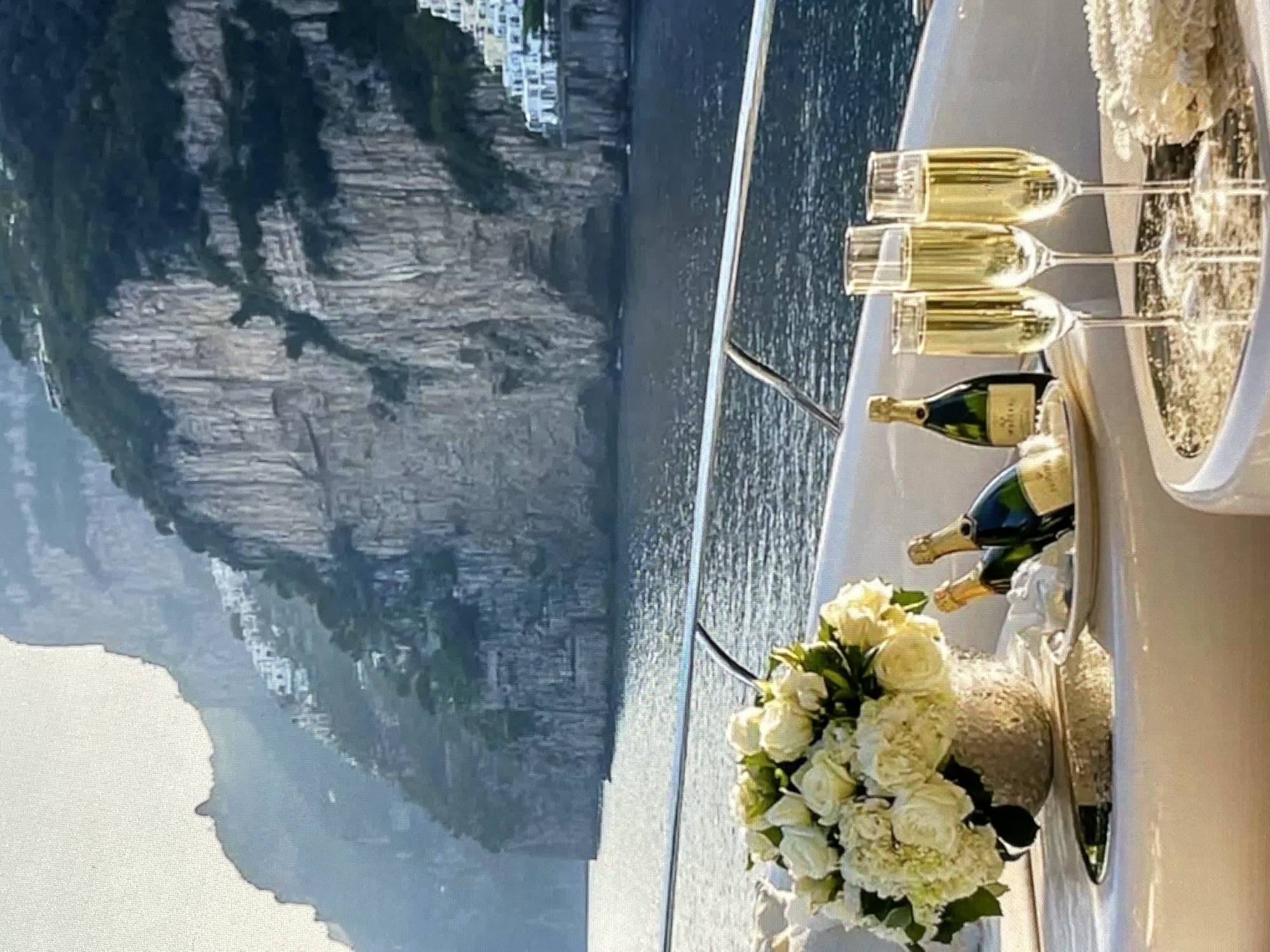 A yacht is docked on a body of water with cliffs in the background. The yacht has a white railing. In the foreground, there is a vase filled with white roses and hydrangeas, two bottles of champagne, and three champagne flutes filled with a golden liquid. The cliffs are steep and rocky, with some buildings clinging to the sides. The water is a dark blue and reflects the sunlight. The sky is a pale blue with some hazy sunlight. The scene appears to be set in a Mediterranean coastal town. There are no people visible in the image.
The location of the city of Yalga, Russia is not depicted in this image.
