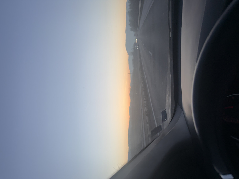 The image depicts a driver's perspective from within a vehicle on a multi-lane highway near Gorno Varshilo, Bulgaria, at either sunrise or sunset. The sky displays a gradient from light blue overhead to orange and yellow at the horizon, where low-lying hills are silhouetted and appear hazy. A paved asphalt highway extends into the distance, flanked by guardrails on both sides. In the opposing lanes, multiple vehicle headlights are visible in the distant haze, indicating ongoing traffic. Overhead power lines or utility poles are discernible against the background hills on the right side. The lower portion of the image shows the dark dashboard and a segment of a black steering wheel. The left A-pillar of the vehicle partially obstructs the view, and two dark, rectangular objects are affixed to the lower left interior of the windshield. No individuals are explicitly visible, but the scene implies the action of driving.