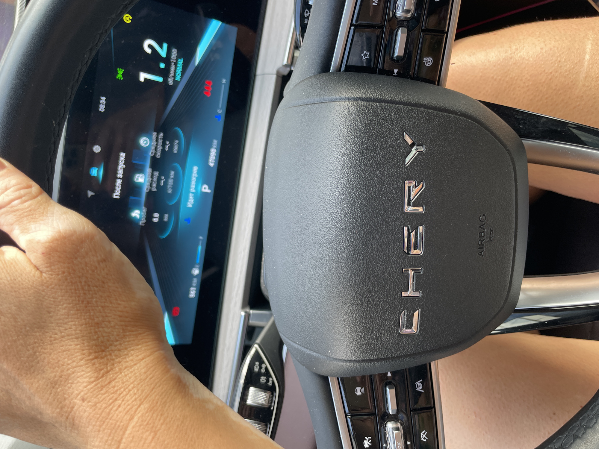 The image shows a close-up of a person's hand gripping a steering wheel inside a Chery vehicle, likely in Sochi, Russia. The vehicle's digital dashboard is visible, displaying information such as "08:34", "1.2 об/мин-1000 NORMAL", and "47090 km". The hand appears to be that of an adult, with visible skin texture. The steering wheel is black leather with chrome accents and features controls on its sides. The interior of the car is modern, with a sleek dashboard design. The lighting suggests it is daytime, with sunlight casting shadows. The background is blurred but appears to be the interior of a car.
