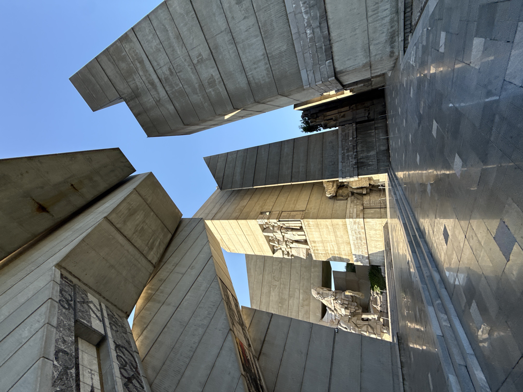 The image shows a low-angle perspective of the Founders of the Bulgarian State Monument in Shumen, Bulgaria. The monument is constructed from large, rectangular concrete blocks. These blocks form abstract shapes against a clear blue sky. Some blocks feature carved depictions of figures. A paved area is visible on the right side of the frame, reflecting light.