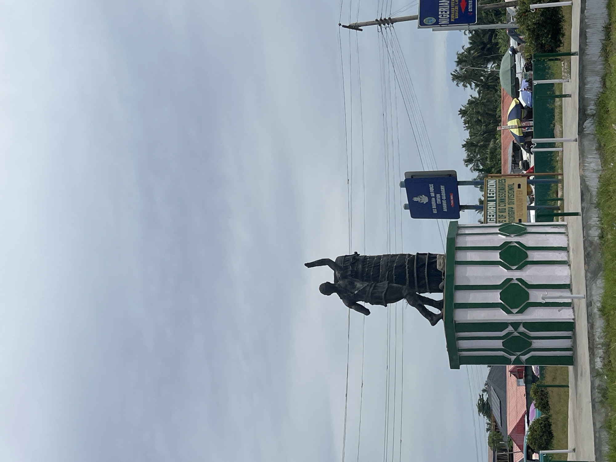 The image shows a statue of a man carrying another man on his back in Badagry, Nigeria. The statue is positioned on a white and green base with geometric patterns. Nearby are signs for the "653 Nigerian Air Force Station Ahanve-Dadagry" and "GERIAN LEGION". Telephone/electrical wires stretch across the top of the image. In the background, there are trees, a glimpse of buildings, and several people.
