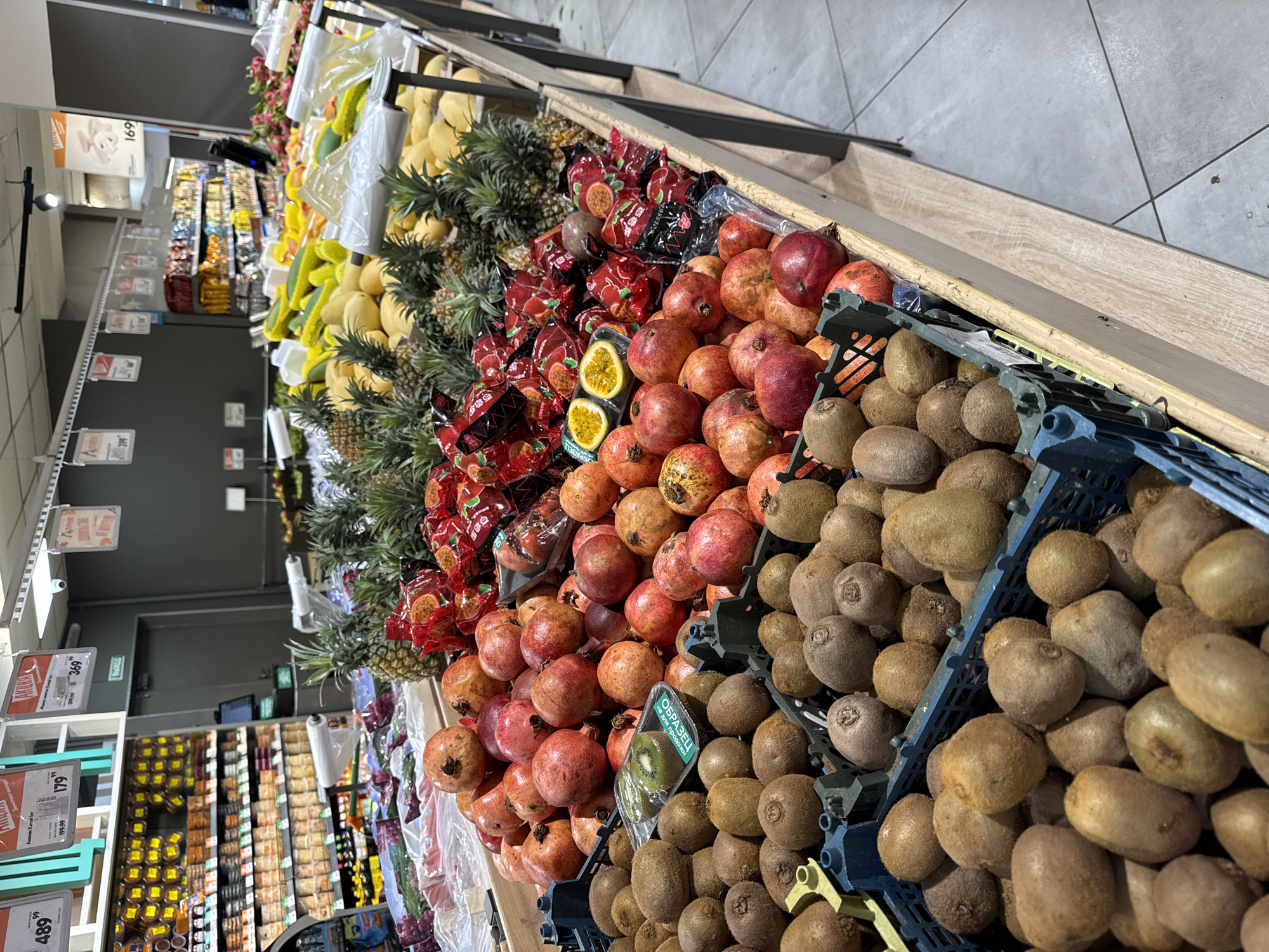 The image shows a fruit display in a supermarket aisle. The location is identified as Krasnoyarsk, Russia (based on Cyrillic text on price tags). The display features several varieties of fruit, arranged in rows: kiwis in blue plastic crates, pomegranates, bagged passion fruit, pineapples, mangoes, and other fruits further back in the display. The aisle is filled with shelves of packaged goods visible in the background. Price tags are visible beneath the fruits. The scene is brightly lit by overhead fluorescent lights. No people are visible.