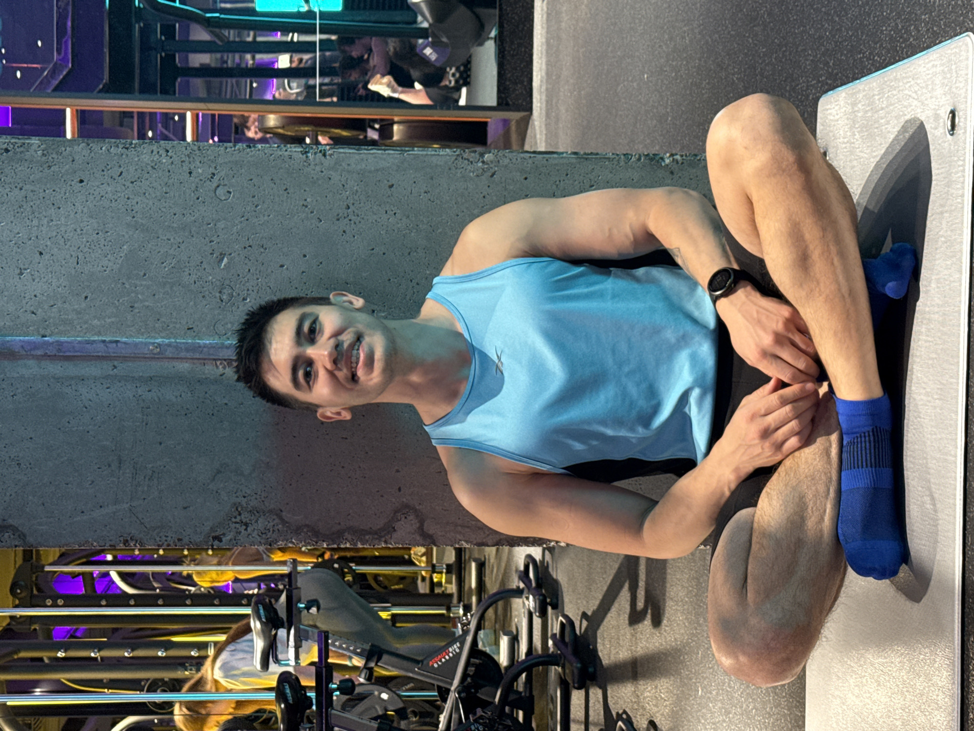 A young man is seated in an indoor gym, smiling and looking directly at the camera. He wears a light blue tank top, black shorts, and royal blue athletic socks, with a black smart watch visible on his left wrist. He is sitting on the floor or a low surface, with one leg bent and the other crossed, possibly on a light grey exercise mat that extends to his right.
Behind him to the left, there is a prominent concrete pillar or wall. The background reveals reflections of gym equipment, likely in a large mirror or glossy surface, showing metallic structures consistent with exercise machines such as a recumbent bike or leg press. Partial text "ASS..." and "CLASSIC" is discernible on this reflected equipment, possibly indicating an "AssaultBike Classic." Vibrant purple and turquoise neon or LED lights are also visible within these reflections, contributing to a modern and dynamic gym ambiance.
The man's direct gaze and cheerful smile suggest a friendly and relaxed demeanor. The setting is clearly a fitness center, characteristic of gyms found in urban environments like Kazan, Russia. The time of day is not explicitly indicated due to the indoor lighting.