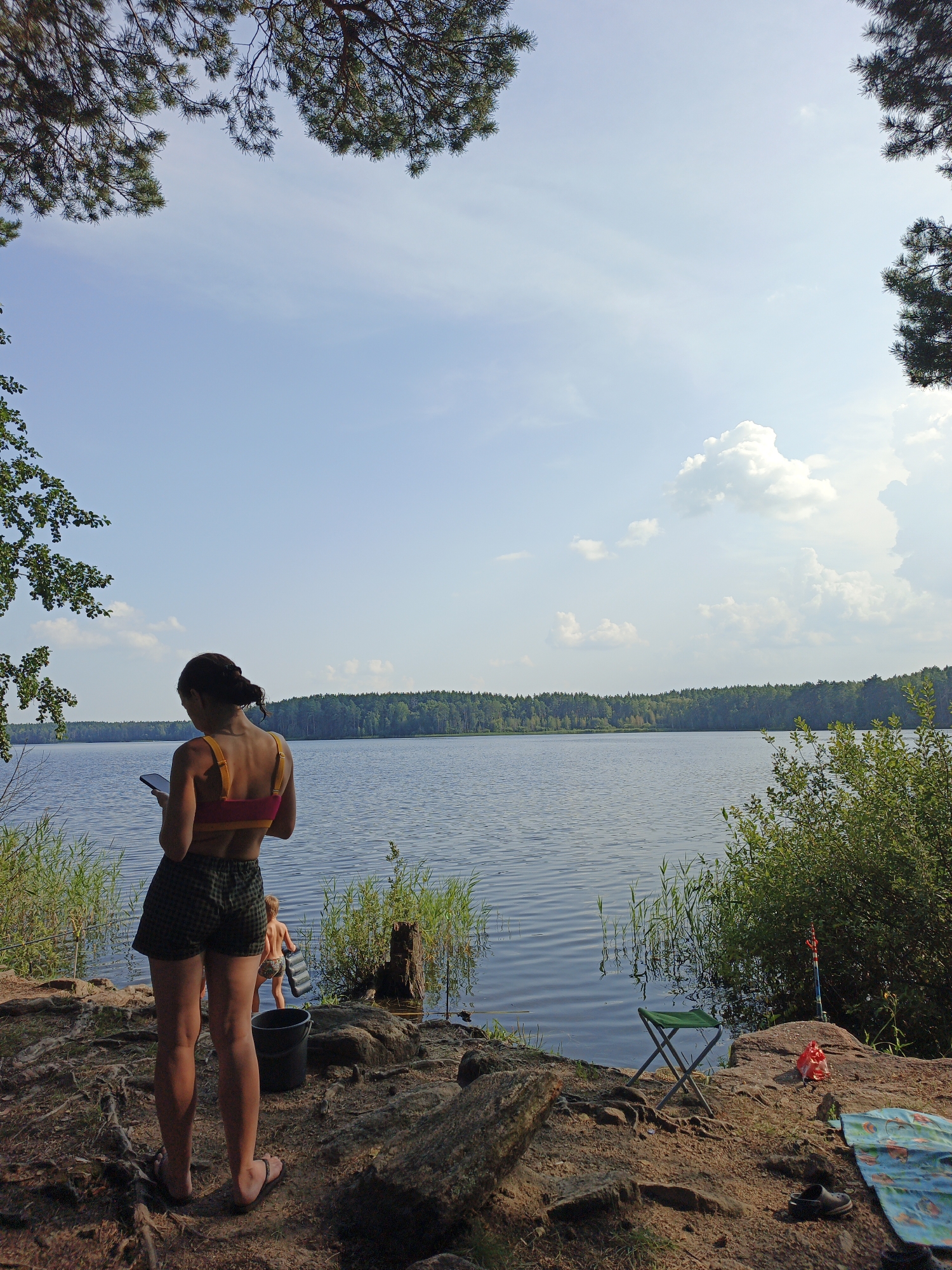 This is a wide, eye-level, outdoor, daytime shot of a lake scene in Sarafanovo, Russia. In the foreground, a young woman stands with her back to the camera, looking at her phone. She is wearing a pink and yellow crop top and checkered shorts. To her left, a black bucket sits on the ground, and to her right, a green folding chair, a red bag, and a blue patterned mat are visible. A fishing rod stands upright in the background. A child, partially submerged in the water, can be seen in the middle ground. The lake stretches across the background, bordered by a dense forest. The sky is blue with scattered clouds. The ground is a mix of dirt, rocks, and tree roots. Pine branches frame the top of the image.