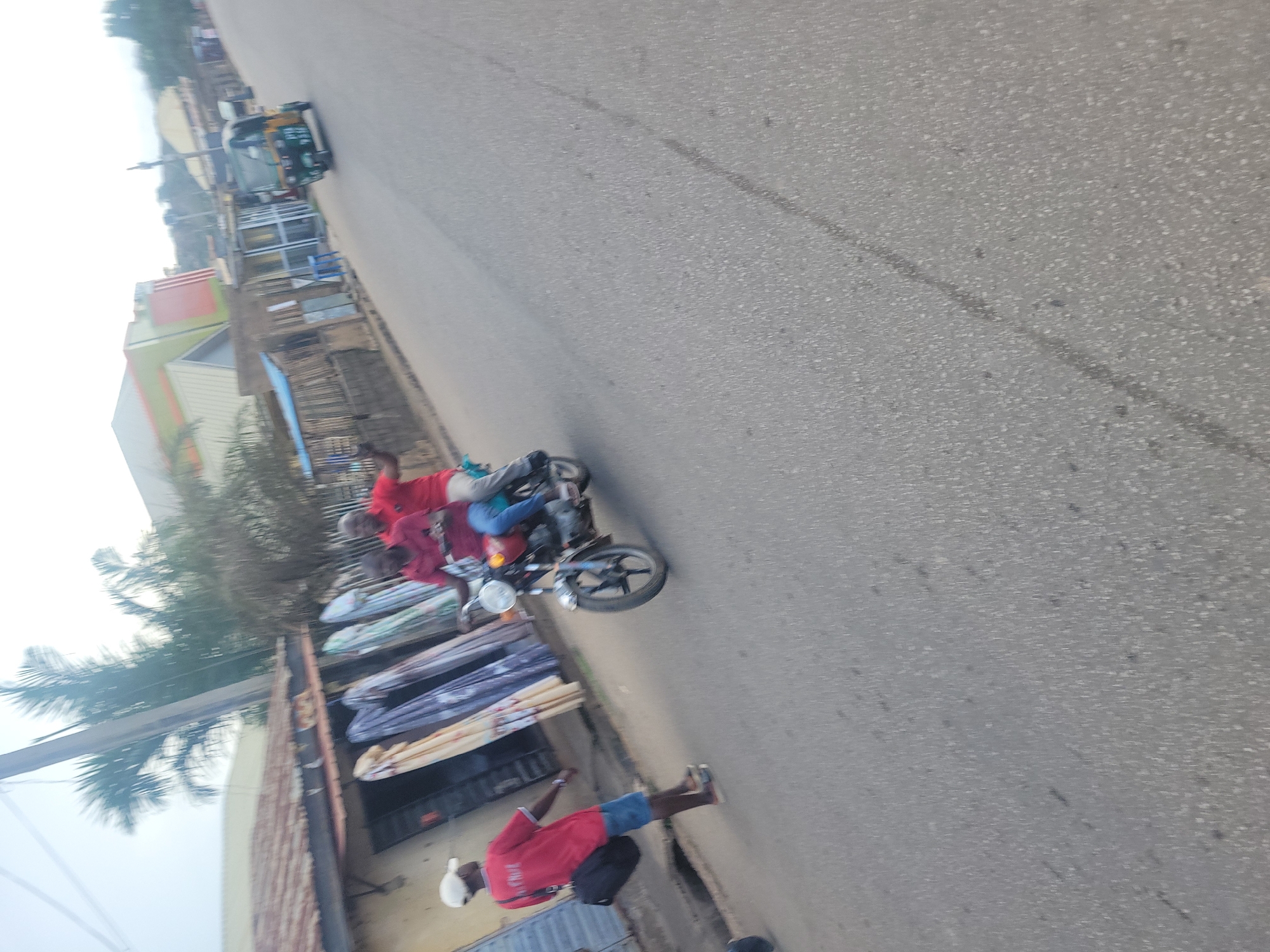 In Jos, Nigeria, a man in a red t-shirt and denim shorts walks on a paved road, carrying a black bag over his shoulder. He is accompanied by two people on a red motorcycle. The motorcycle is being ridden by a man in a red shirt and jeans, with another person in a red shirt behind him. A yellow and green tricycle is visible in the background, parked on the side of the road. The scene appears to be a marketplace with shops and stalls lining the street, selling fabrics. The sky is overcast.