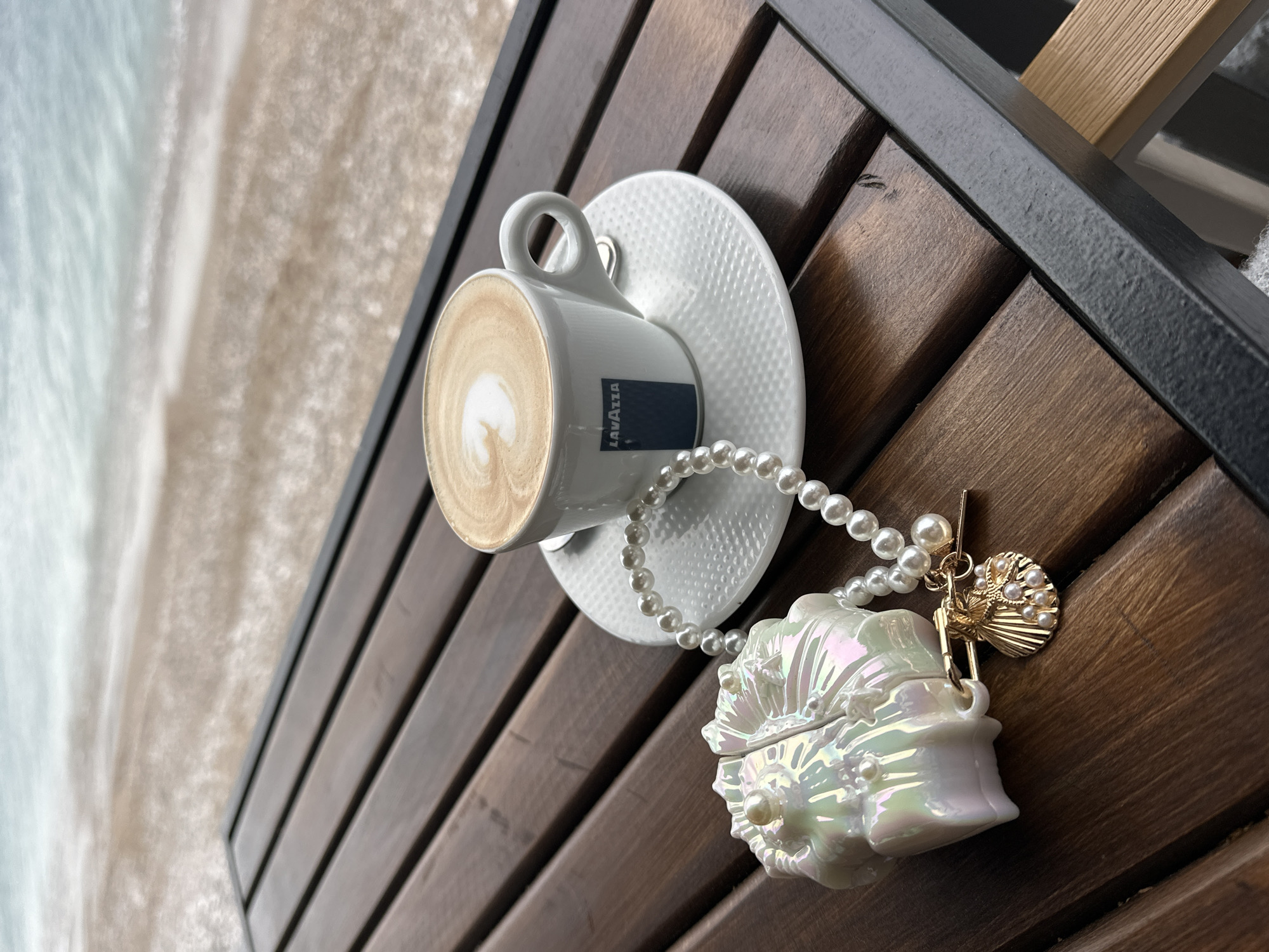 A top-down, close-up shot shows a Lavazza cappuccino on a white saucer, a string of pearls, and a shell-shaped jewelry box with a gold charm on a wooden table. The cappuccino features latte art, and the pearl necklace drapes from the saucer to the jewelry box. In the upper left corner, the blurred coastline of Šušanj, Montenegro, is visible. The table has dark wooden slats, and part of a metal frame is visible along the top.