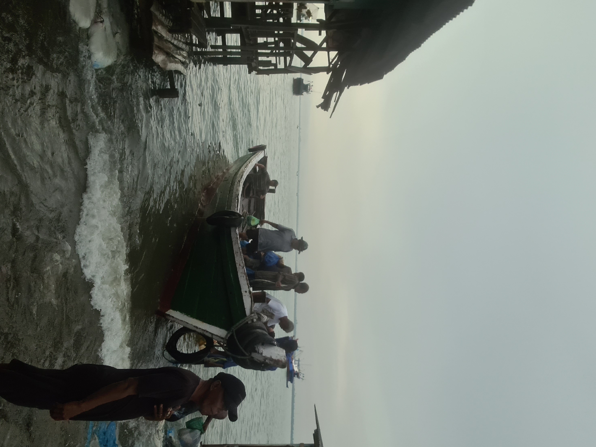 This is a medium-angle, outdoor, full-shot image taken at the Lhokseumawe fish market, Indonesia. The sky is overcast, with a light gray hue. In the foreground, a man in a black t-shirt and black cap stands facing right, his hand resting on his chest. Beside him, to the left, is a traditional fishing boat with a green hull and white trim, partially submerged in the water. Several people are on board the boat, some appearing to be loading or unloading goods. The boat is docked near a wooden structure on stilts, which appears to be part of the market. In the background, several other fishing boats and a larger vessel are visible on the water. The sea appears calm with small waves lapping at the shore. The scene captures a typical day at the bustling fish market.