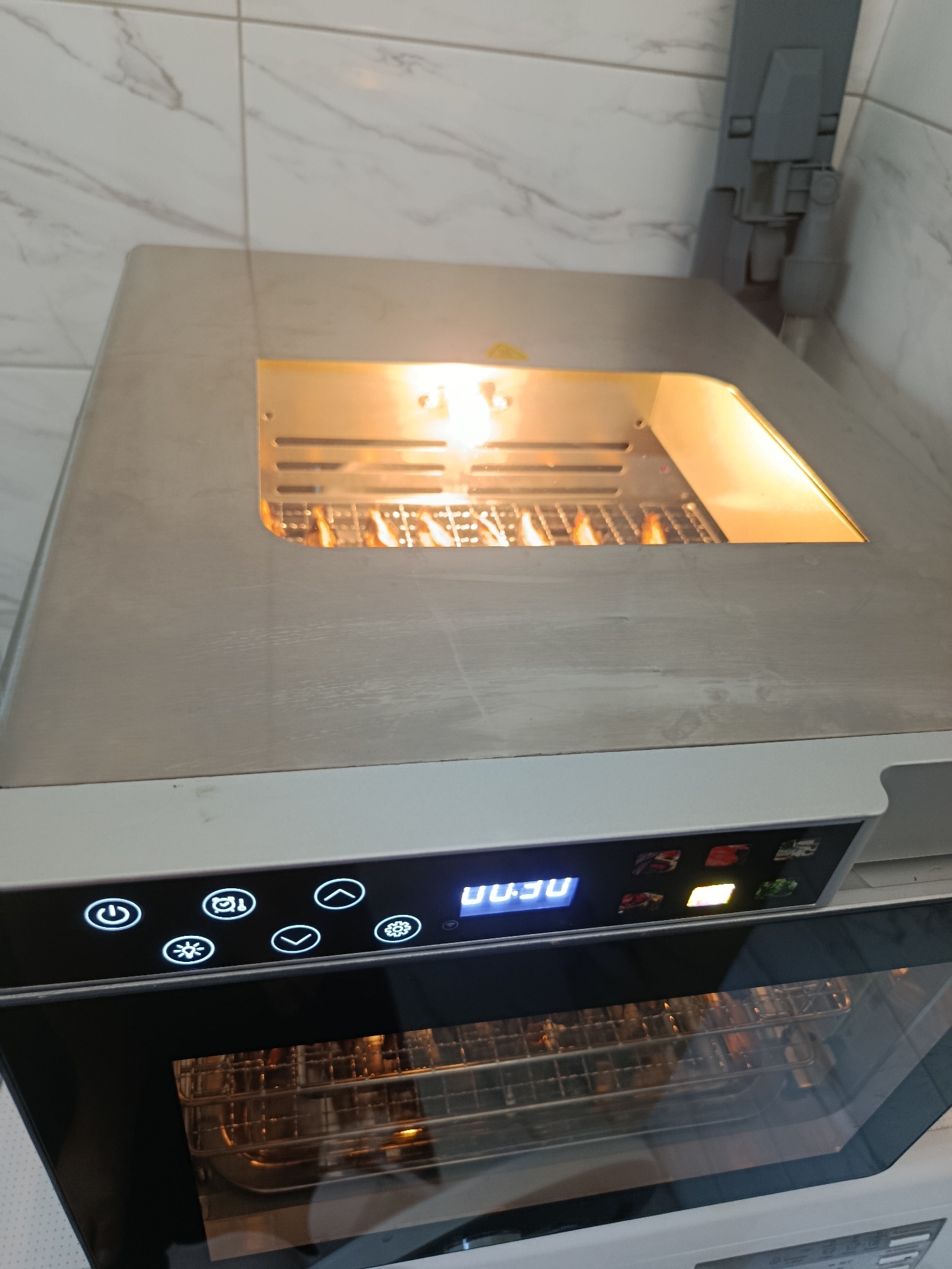 An indoor close-up shot shows the top of a modern, silver and white oven. The oven has a digital display panel with illuminated icons and blue digits reading "00:30". Above the panel, a stainless steel panel has a rectangular opening revealing a wire rack with food items on it, illuminated by internal heating elements that glow orange. To the right of the oven's top, a grey plastic appliance, possibly a vacuum cleaner hose attachment or a part of the kitchen ventilation system, is attached to the wall. The wall is tiled with a white marble-patterned ceramic material. The overall environment suggests a kitchen setting in Vyselki, Russia.