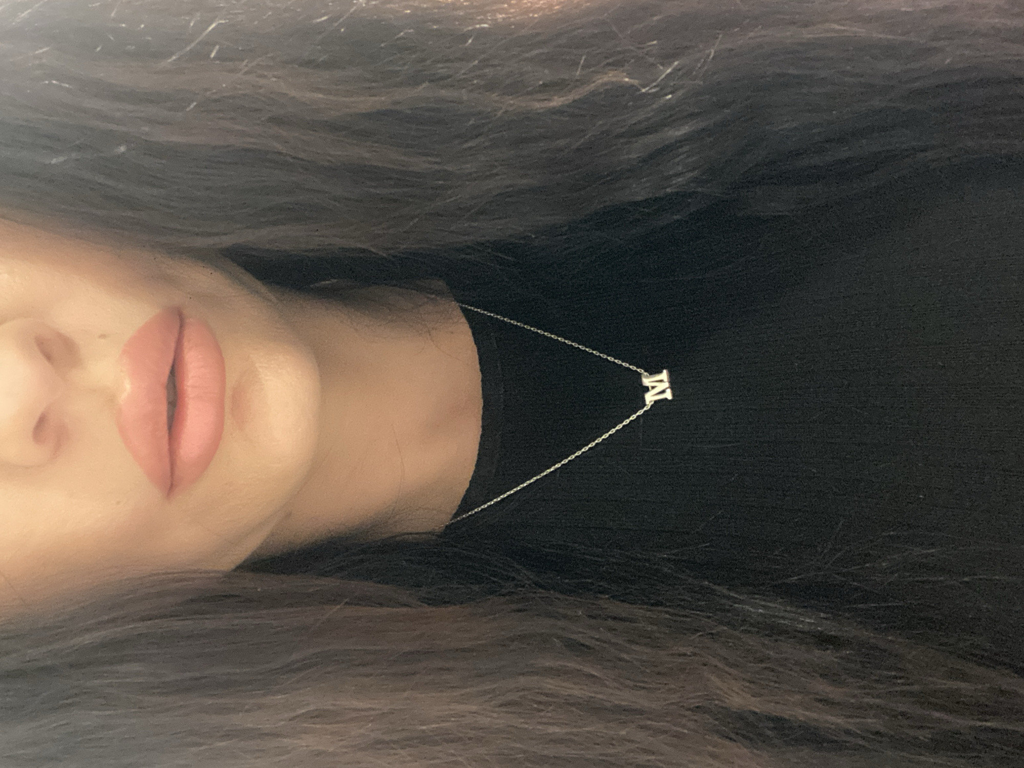 A close-up, chest-up, medium shot captures a person with dark brown hair parted down the middle and framing their face. The person is wearing a black, ribbed, long-sleeved shirt. A delicate silver chain necklace with a single silver letter "M" pendant rests on the shirt. The person's lips, painted with a soft pink lipstick, are slightly parted, suggesting a neutral or slightly open expression. The background is dark and undifferentiated. The image does not contain any contextual information that would allow for the determination of its location in Sharm El Sheikh, Egypt.