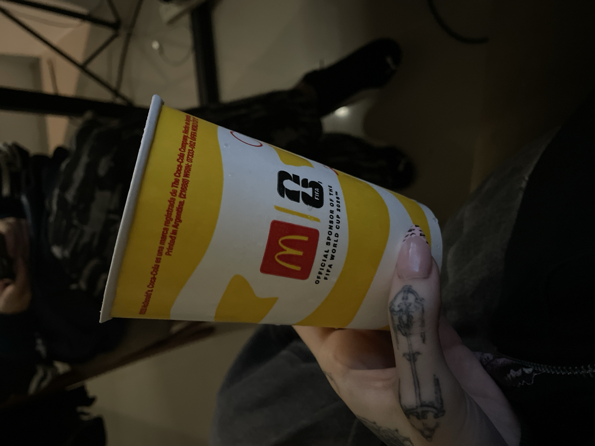 A person's hand, adorned with light pink nails featuring black French tips and a detailed tattoo of an ornate key-like design on the middle finger, holds a McDonald's paper cup. The cup, decorated with yellow and white wavy stripes, prominently displays the red McDonald's "M" logo. Below it, the FIFA logo is visible, accompanied by a stylized "26" and text stating "OFFICIAL SPONSOR OF THE FIFA WORLD CUP 2026™."
Further text on the cup includes Spanish phrases: "McDonald's, Coca-Cola es una marca registrada de The Coca-Cola Company. Hecho en Argentina. Printed in Argentina." This text, along with numerical codes like "(21688) WRIN: 07333-062," firmly places the context in Argentina, consistent with Buenos Aires.
The scene is dimly lit, suggesting either nighttime or a dark indoor environment. The background is out of focus, showing dark fabric, likely the person's clothing (such as a lap or legs), and indistinct shapes that could be furniture or other elements of a casual indoor setting. The primary activity depicted is simply holding the beverage cup. No specific emotions are discernible.