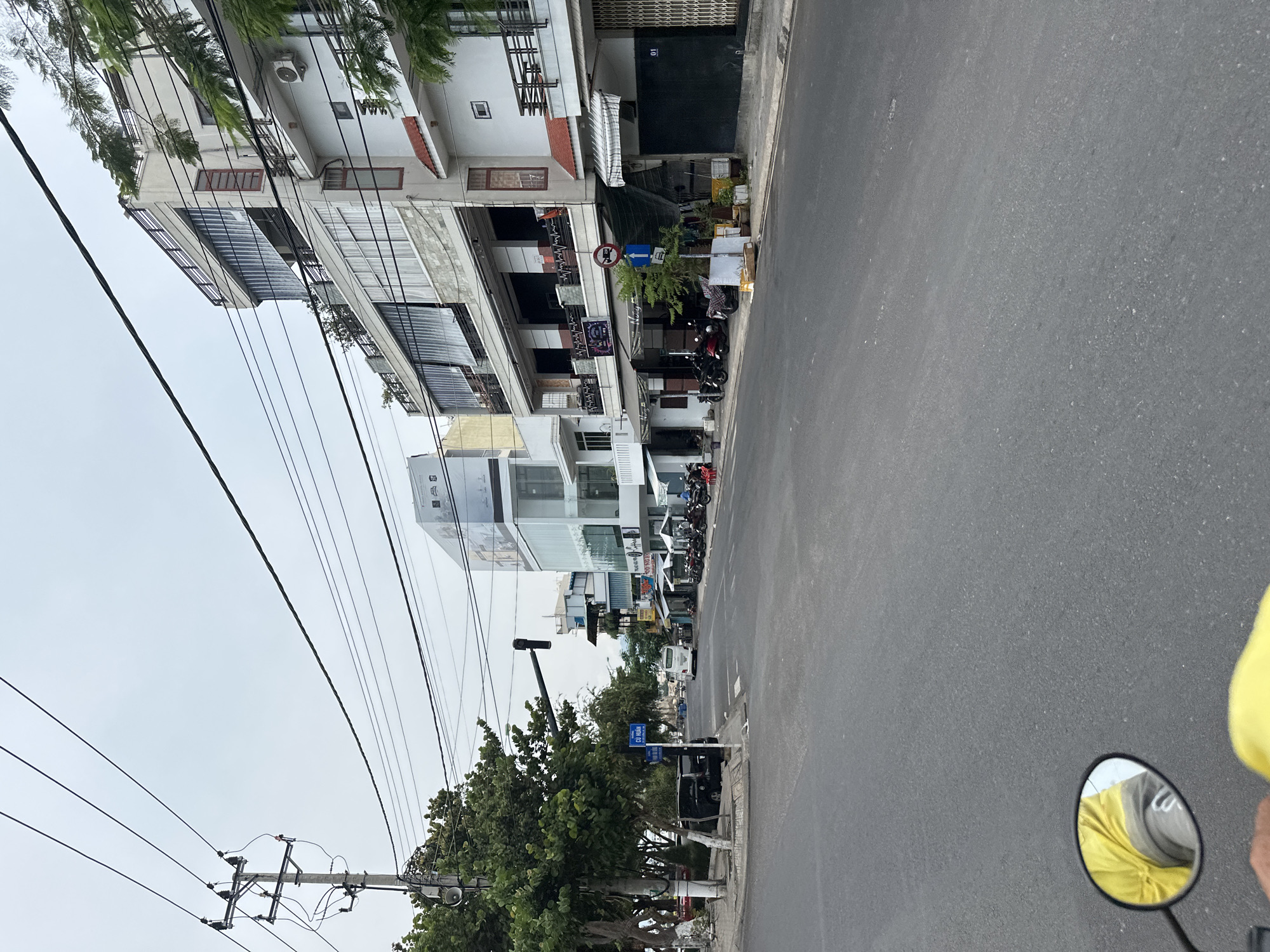 The image shows a street scene in Nha Trang Ward, Vietnam. The view is looking down a street lined with multi-story buildings. A row of buildings is on the right of the image with a variety of architectural styles. Many motorbikes are parked alongside the buildings. The road is paved with asphalt. Overhead power lines traverse the sky. Trees and street signs are on the left side of the road. In the bottom right, a portion of a rearview mirror from a motorbike is visible. The sky is overcast.