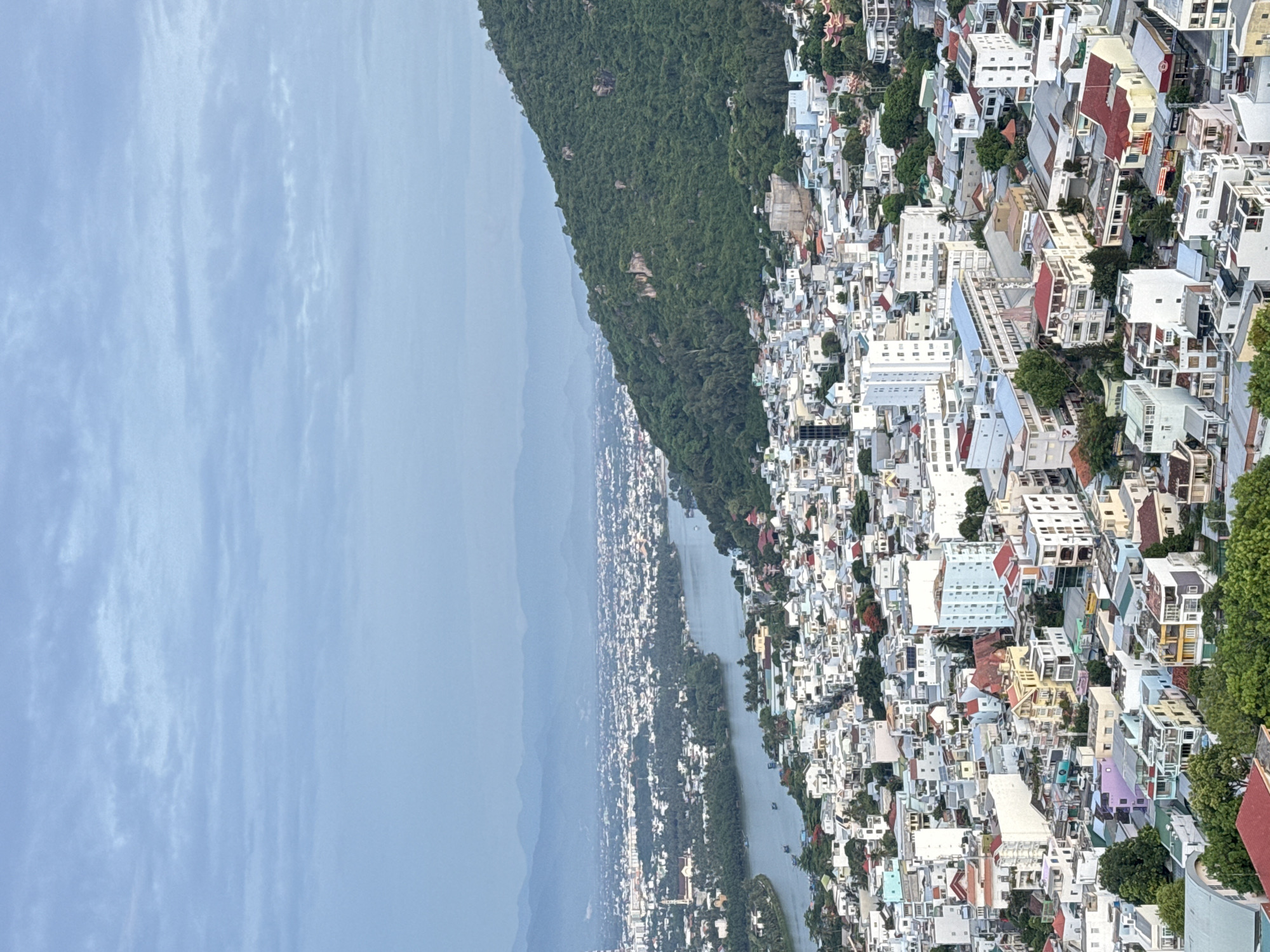 This is a high-angle aerial view of a densely populated city in Nha Trang, Vietnam, with a wide river flowing through it. The city is characterized by a multitude of low-rise buildings, many painted in white, with terracotta tile roofs. Some buildings are taller and more modern, featuring concrete facades and balconies. The architecture suggests a mix of residential and commercial structures.
The river, appearing calm and a light gray color, meanders from the background towards the foreground, with a few small boats visible on its surface. On the right side of the image, a large, verdant hill covered in dense green trees slopes down towards the city, its rocky outcrops partially visible. The background features layers of hazy mountains, receding into a pale blue sky filled with soft, diffused clouds. The overall lighting suggests it is either early morning or late afternoon, with a muted, overcast sky. There are no people or animals clearly discernible in the scene. The scene captures a sprawling urban landscape integrated with natural elements of water and mountainous terrain.