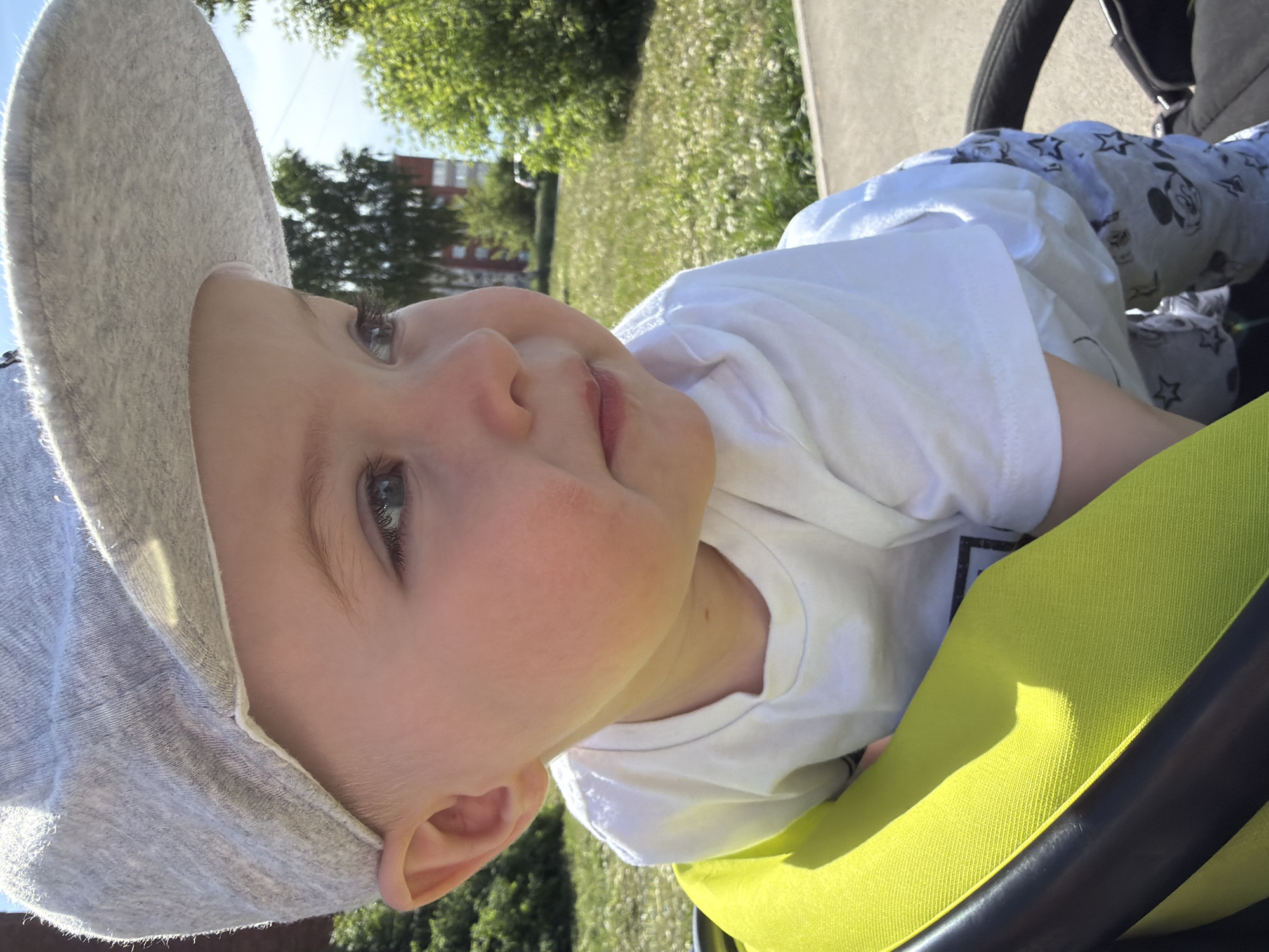A baby wearing a gray sun hat and a white t-shirt is lying in a stroller with a bright green liner and Mickey Mouse-patterned pants. The stroller is on a paved surface next to a grassy area with trees, possibly a park in Votkinsk, Russia. A building is visible in the background.