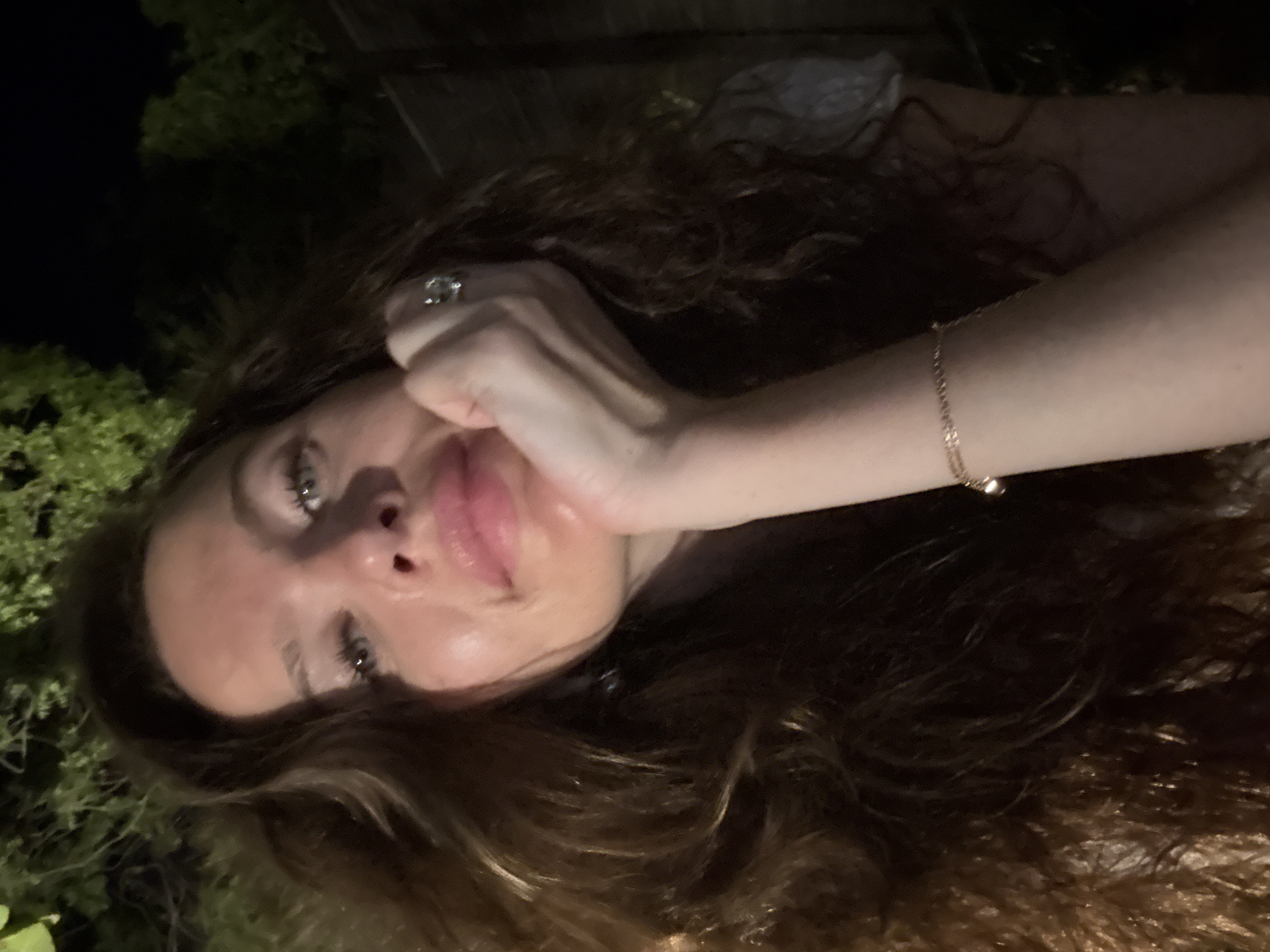 The image shows a close-up of a woman's face, taken at night. She has long, wavy brown hair and is wearing makeup. Her right hand is resting on her chin, and she is wearing a ring on her index finger and a bracelet on her wrist. The background is dark and appears to be outdoors, with some greenery visible. There is no indication of a specific location in Dubai, United Arab Emirates.