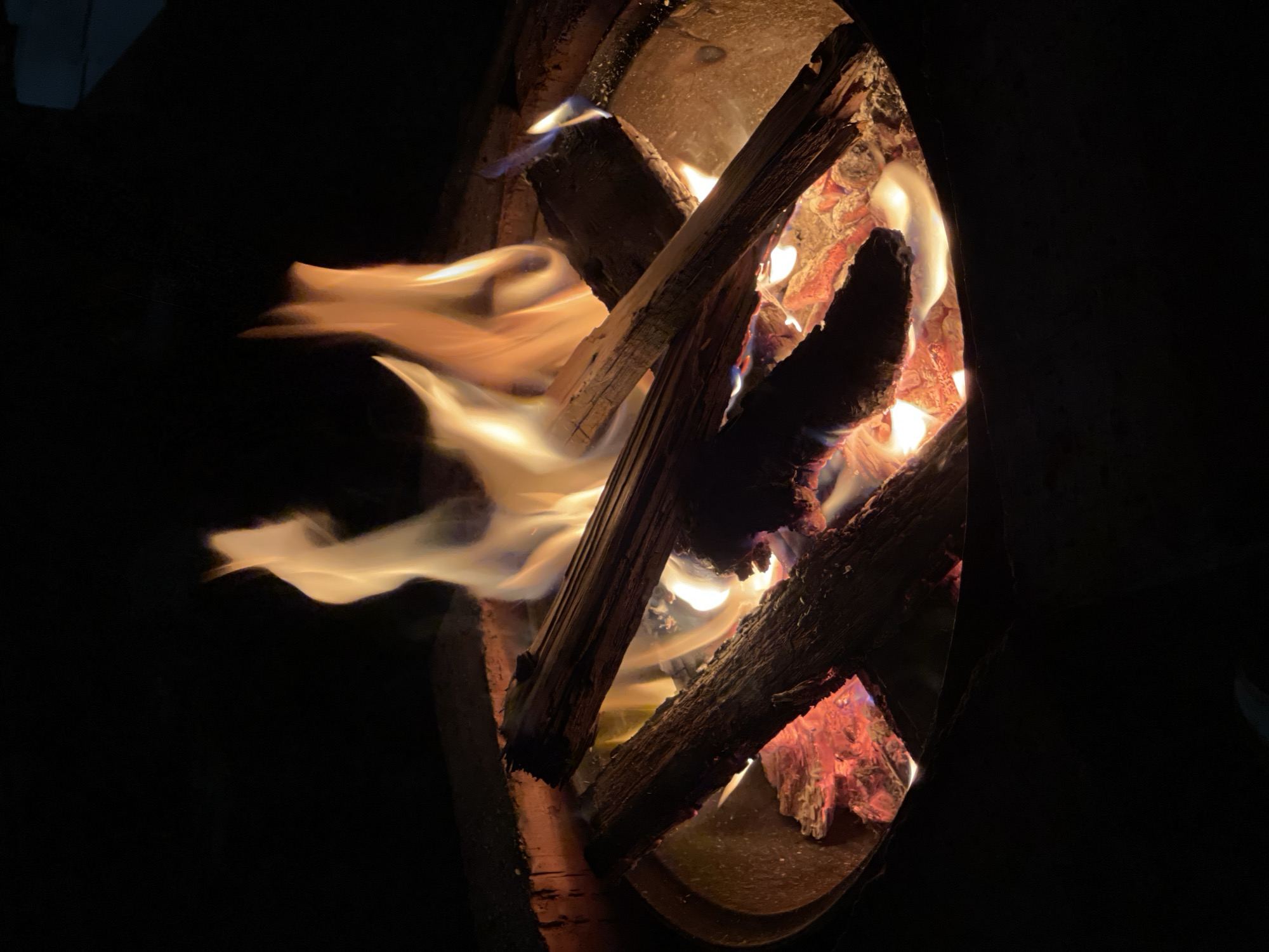 The image shows a close-up of a bonfire at night. The fire is contained within a circular fire pit made of brick or stone. Several pieces of wood are burning, with flames and embers visible. The flames are predominantly yellow and orange, with some white and blue at the base. The background is dark, indicating it is nighttime. There are no people visible in the image. The overall scene depicts a warm and inviting campfire.