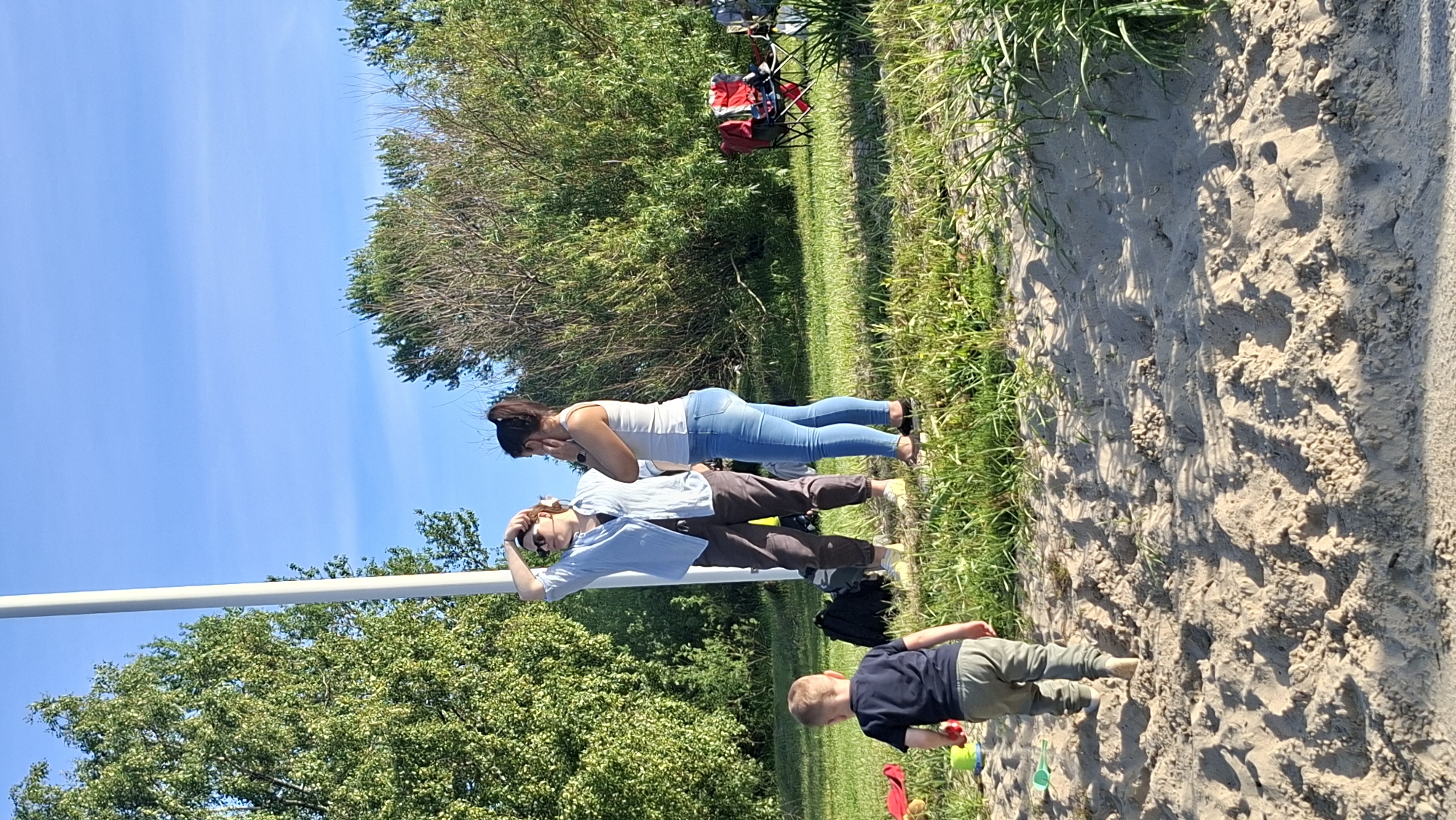 In a sunlit outdoor scene, a young boy clad in a dark t-shirt and olive-colored pants walks away from the viewer, his back to the camera, on a sandy terrain. He is heading towards a patch of grass where two adult women are engaged in conversation. The woman on the left, wearing a light blue button-down shirt, dark pants, and sunglasses, has her hand raised to her forehead. The woman on the right, in a white tank top and blue jeans, is looking away, her hand near her ear. Behind them, a tall white pole stands vertically, with lush green trees and dense foliage forming a natural backdrop. In the background, two foldable chairs, one red and one black, are set up on the grass. The ground is covered with a mixture of sand and grass, with sparse vegetation visible. The sky above is clear and blue. The scene appears to be a public park or recreational area, possibly along the coast of Tallinn, Estonia, given the presence of sandy areas and trees.