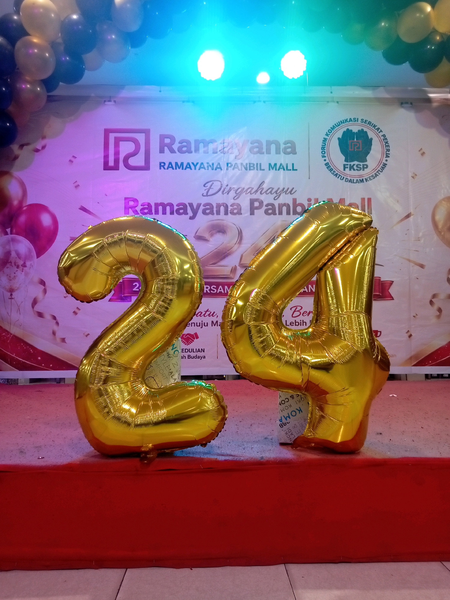In Batam, Indonesia, at the Ramayana Panbil Mall, two large gold foil balloons shaped like the numbers "2" and "4" are displayed on a red stage. Behind the balloons, a banner reads "Ramayana" and "Dirgahayu Ramayana Panbil Mall," suggesting a celebration for the mall's 24th anniversary. The backdrop also features balloons, lighting, and a circular logo with text. The scene appears to be a public event or promotional display.