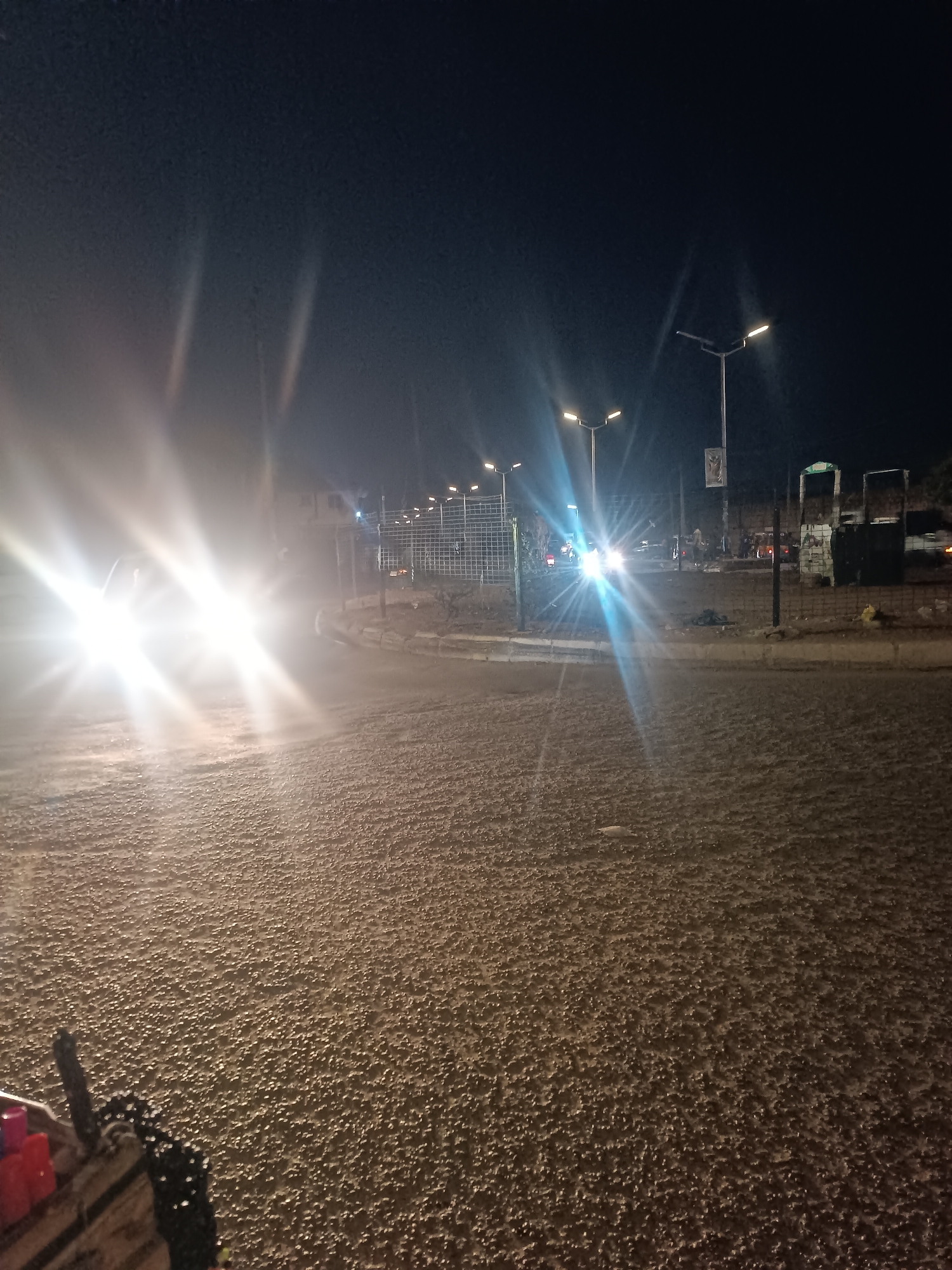 The image depicts a night scene in Bukuru, Nigeria. The foreground shows a paved road surface with a rough texture. Bright headlights from oncoming vehicles are visible on the left side of the image, creating glare. In the background, streetlights illuminate a roundabout. Fences and structures are visible near the roundabout, and other vehicles are present. The sky is dark.