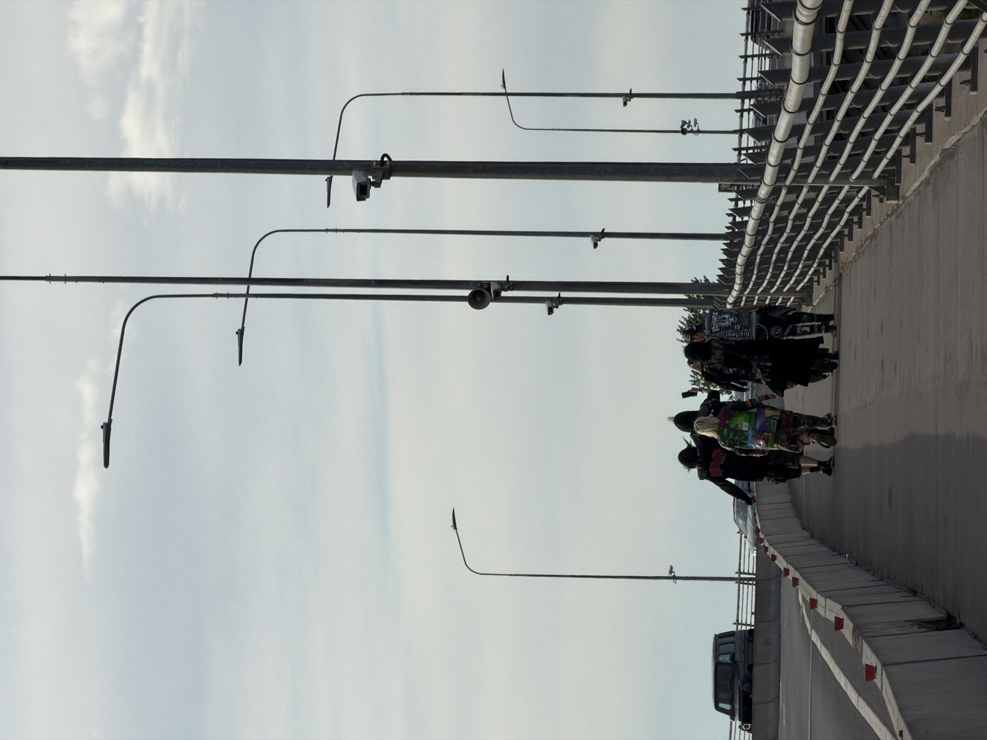 The image depicts a group of people walking on an elevated structure, likely a bridge or overpass, in Saint Petersburg, Russia, under a bright, overcast sky. The group is situated on the right side of the frame, moving towards the viewer's left. Several individuals exhibit distinctive, possibly alternative, fashion styles. One person notably wears a multi-colored, patchwork-style jacket, while another's dark garment clearly displays the white word "DISCLOSE" on their back. They are on a paved walkway, separated from the road by a concrete barrier featuring small red markers along its top edge. A dark-colored truck is partially visible on the road below the walkway. Above the people, multiple dark metal poles extend across the upper portion of the frame. These poles support various devices, including several cameras or sensors, and a speaker-like object, along with light fixtures. On the far right, a complex metallic structure, possibly part of the bridge's support system, is visible, characterized by numerous parallel conduits or pipes. The diffuse lighting indicates it is daytime.